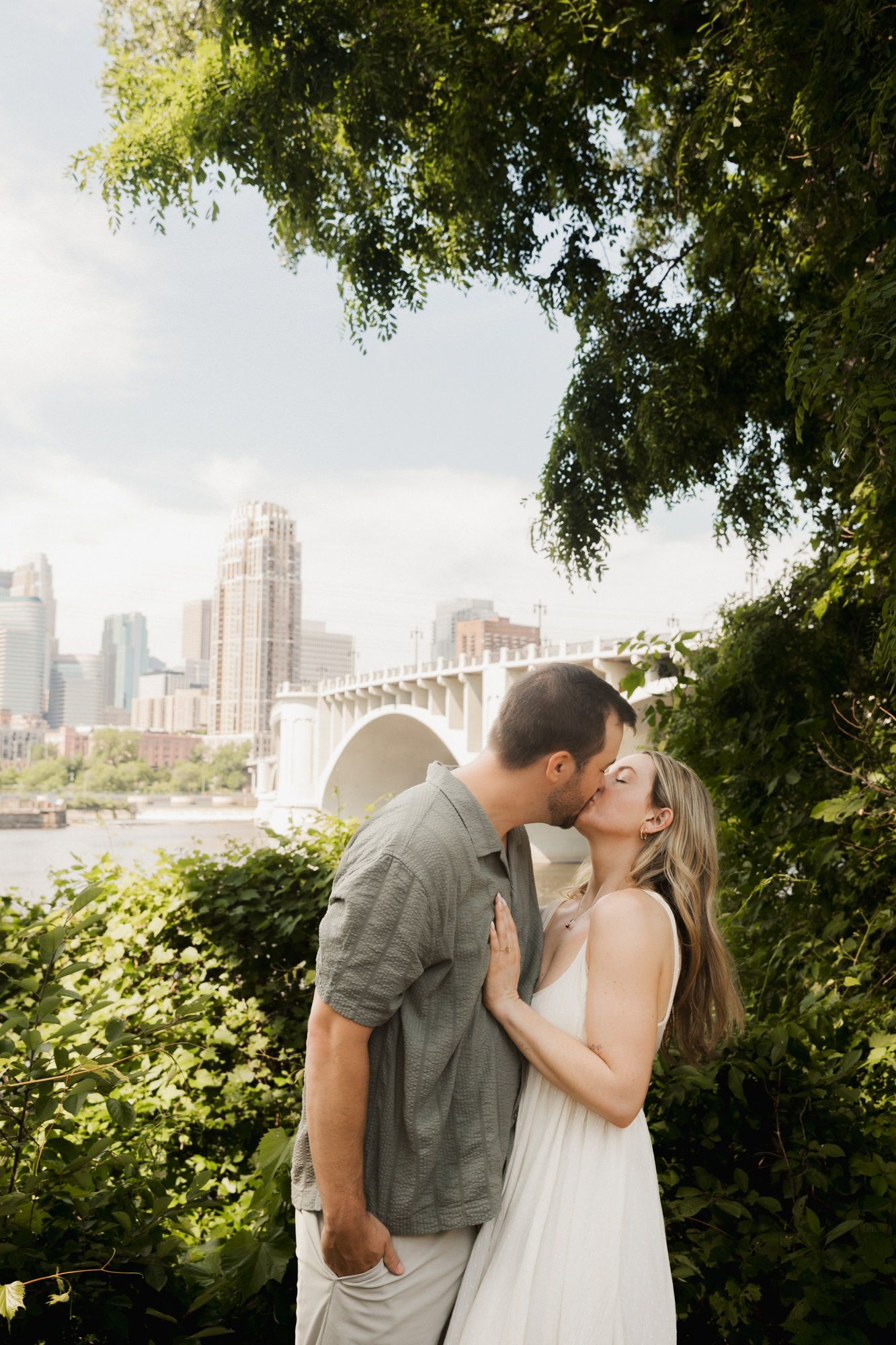 Couple kissing among lush greenery with a city skyline and bridge in the background on a cloudy day.