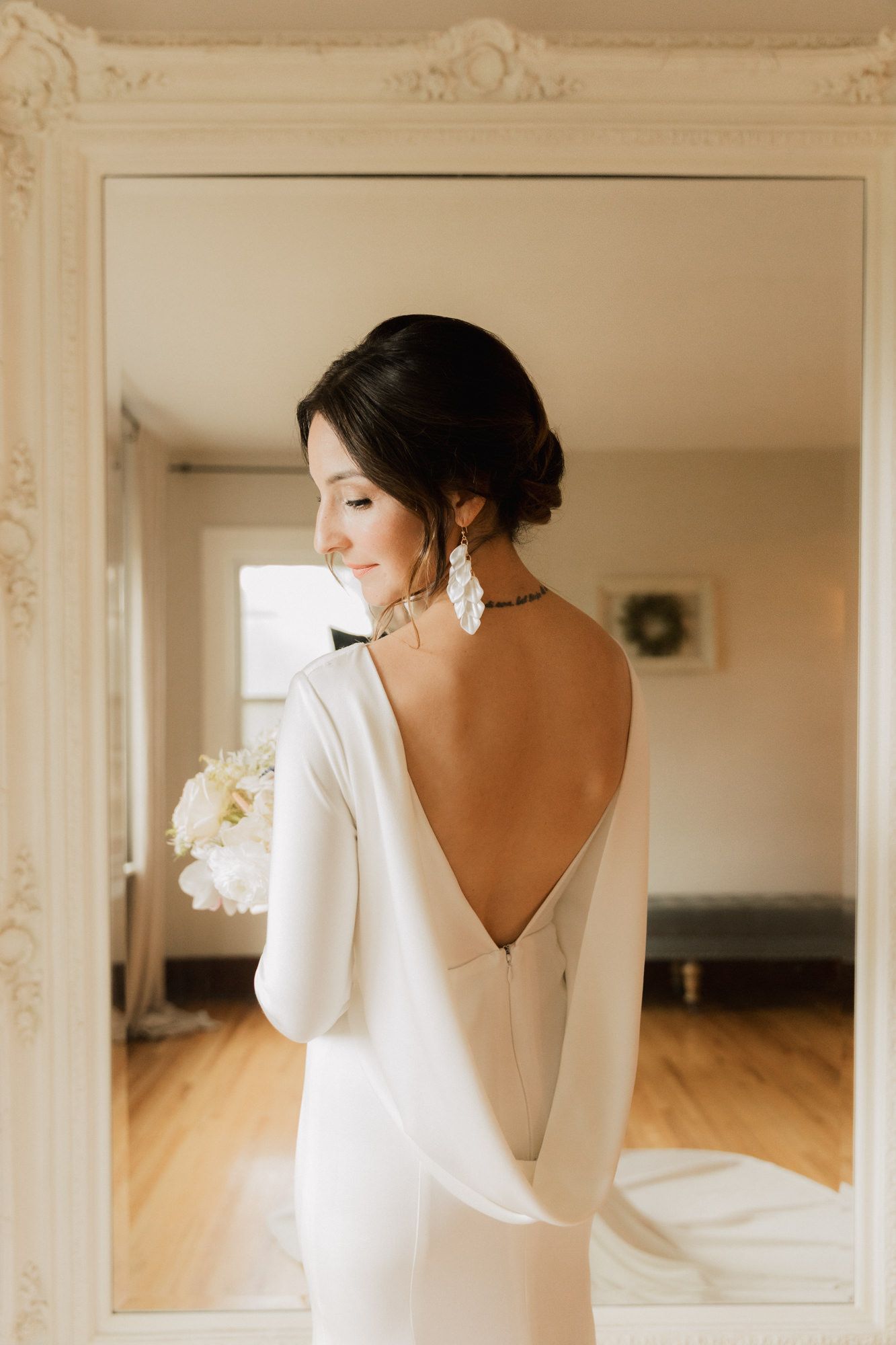 Bride in elegant backless white gown with long sleeves holds bouquet, smiling softly in mirror reflection.