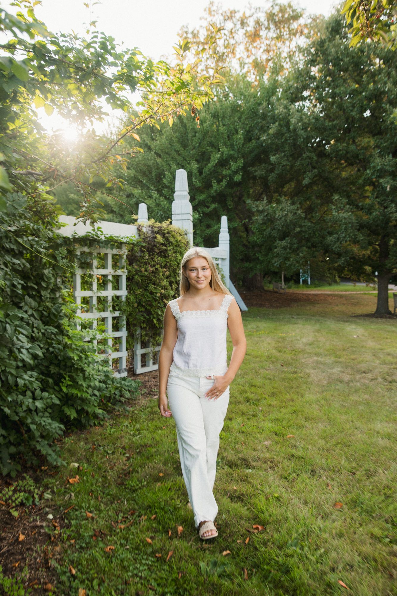 Young blonde woman in white outfit walking through a lush garden with white trellis arbor in sunlight.