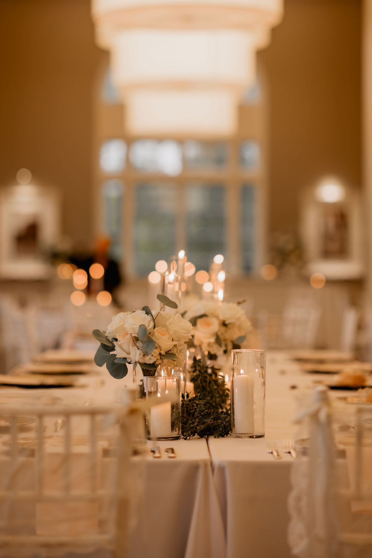 Elegant wedding reception table with white floral centerpieces, candles, and eucalyptus in a warmly lit venue.