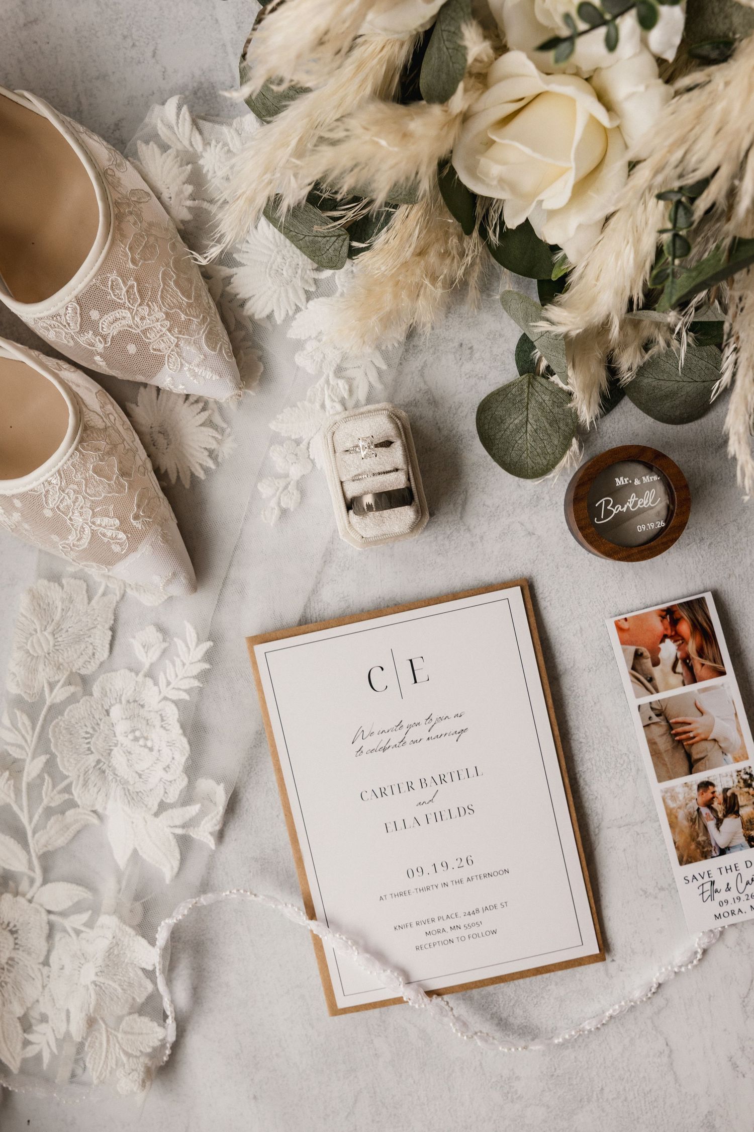 Elegant wedding flat lay featuring lace bridal shoes, white rose bouquet, gold-trimmed invitation, rings, and pampas grass.