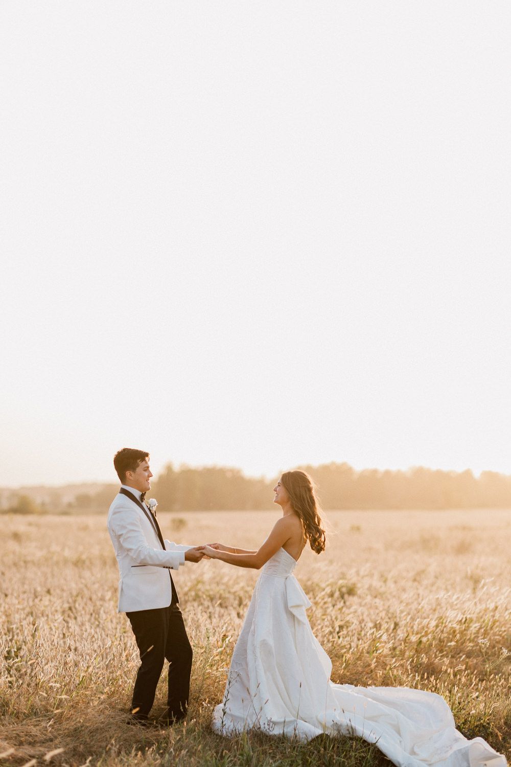 Couple in wedding attire holding hands and dancing in a rustic field at golden hour.