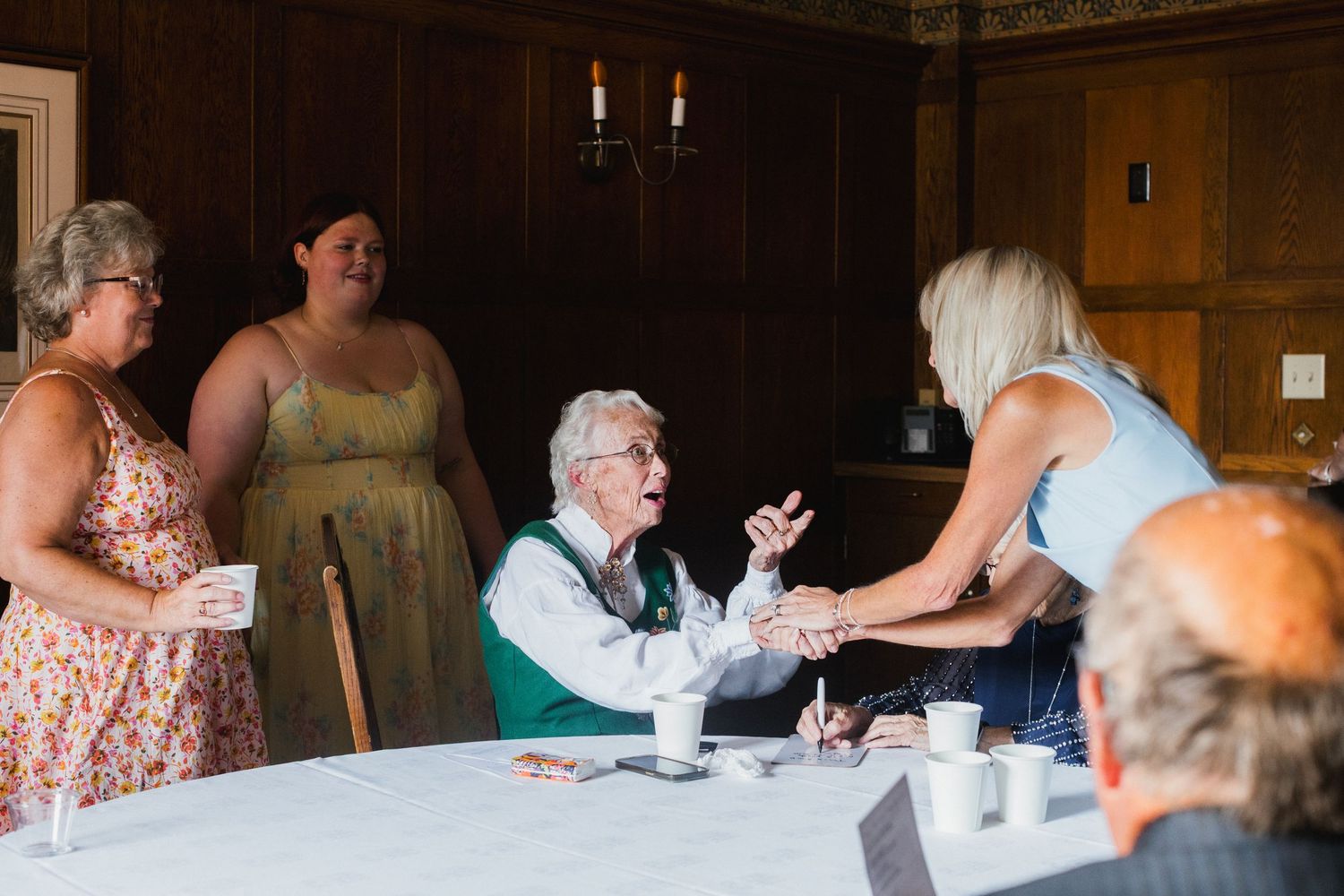 Elderly woman in green jacket conversing with guests at a wedding reception table in a wood-paneled venue.