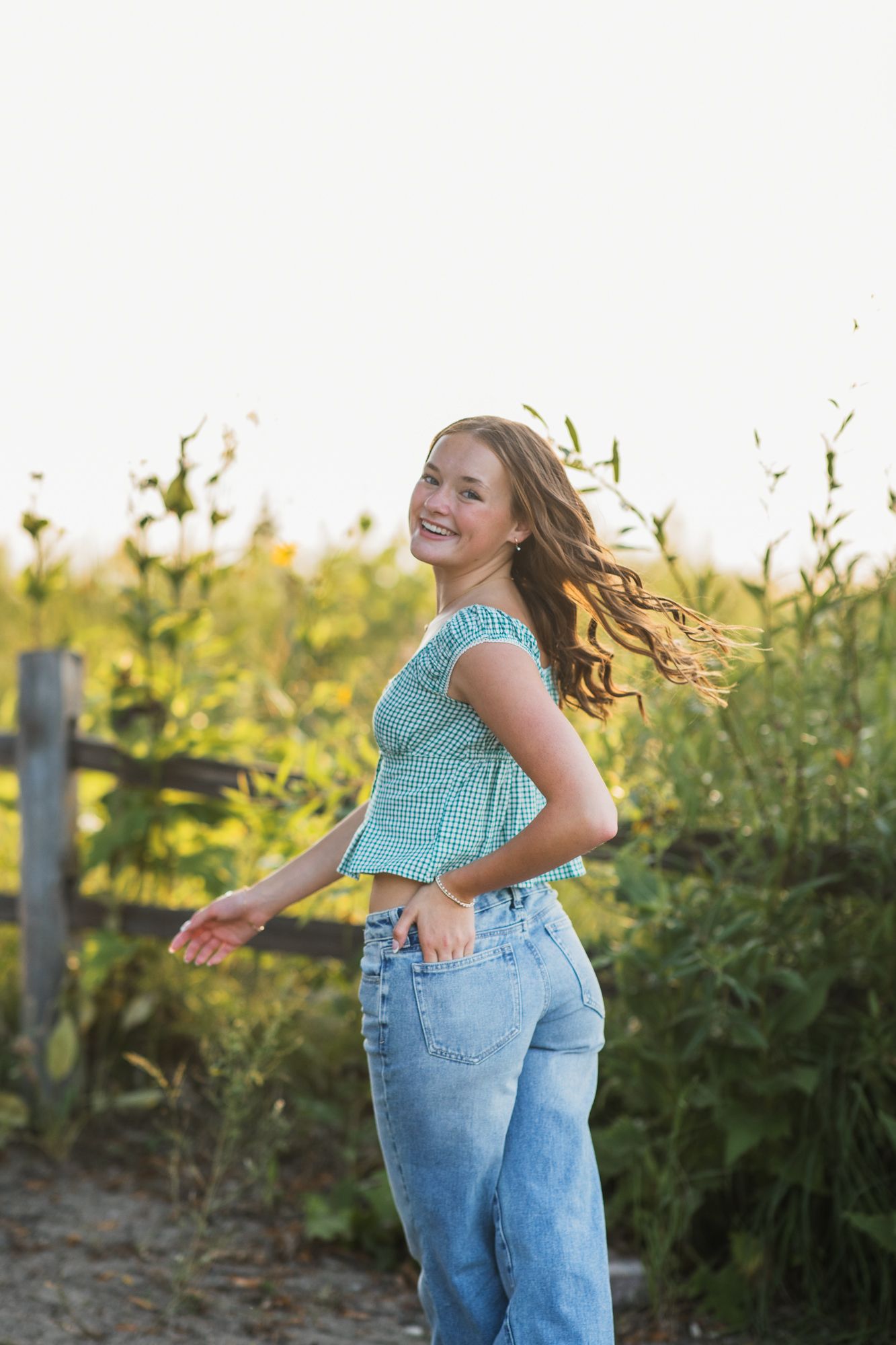 Young woman in teal top and jeans smiling outdoors near sunflowers and wooden fence on a sunny day.