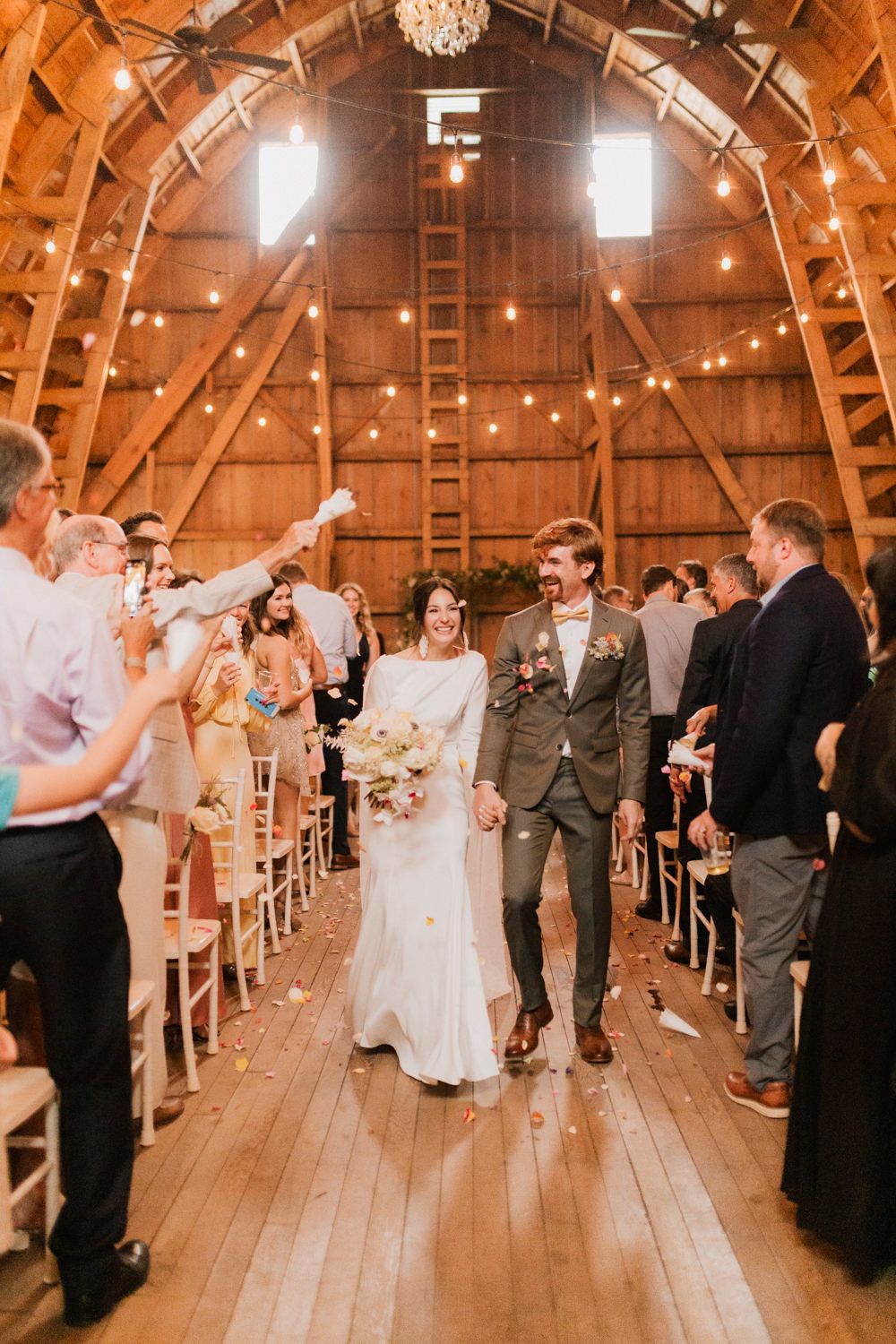 Wedding guests gather in a beautifully lit barn venue with string lights and wooden beams overhead.