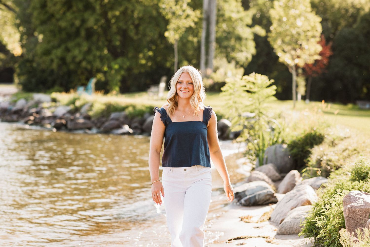 A person in a navy blue dress walks along a sunny lakeside shore with rocks and trees in the background.