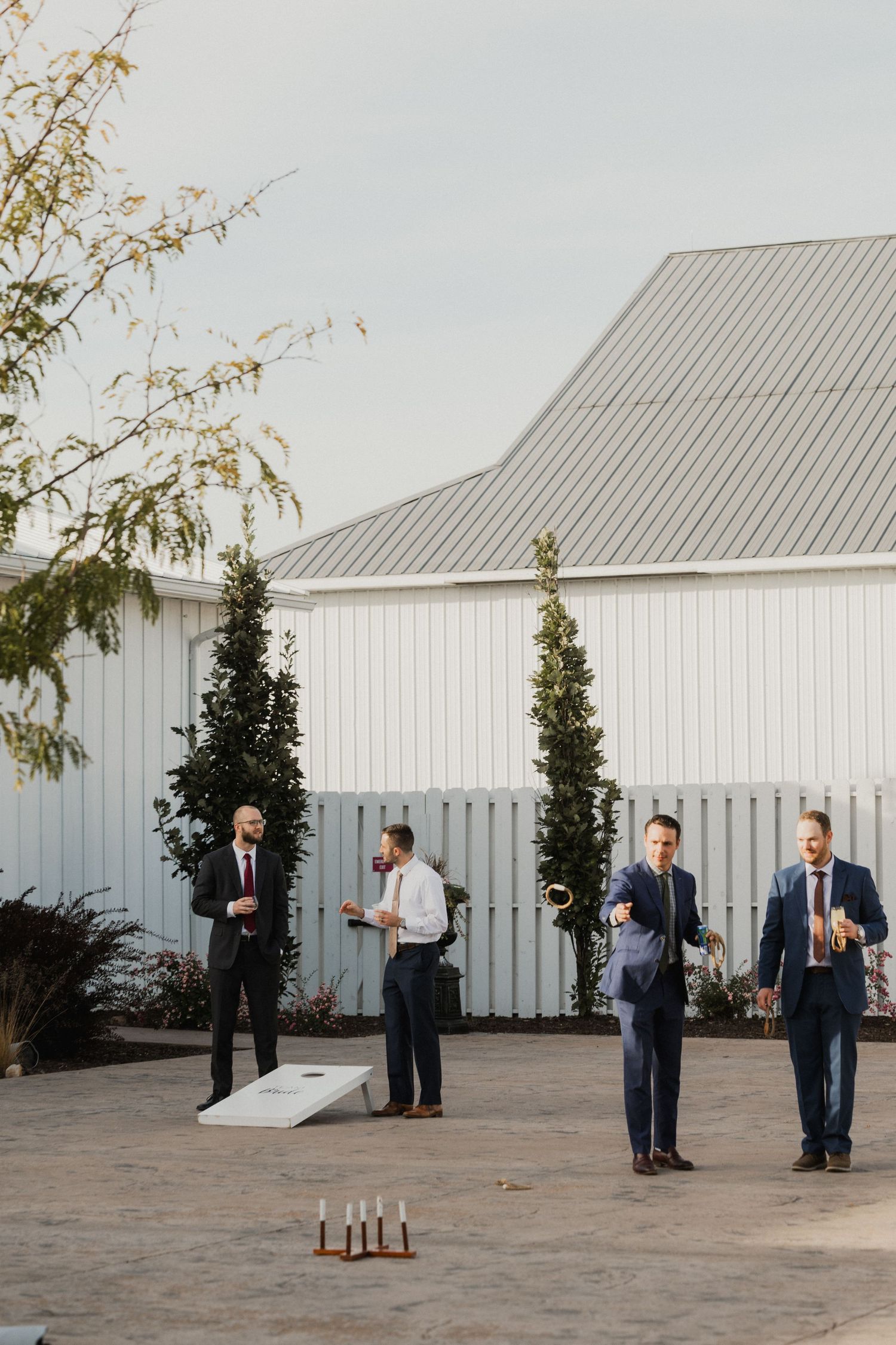 Groomsmen in suits play cornhole and ring toss on a stone patio outside a white barn venue during a wedding celebration.