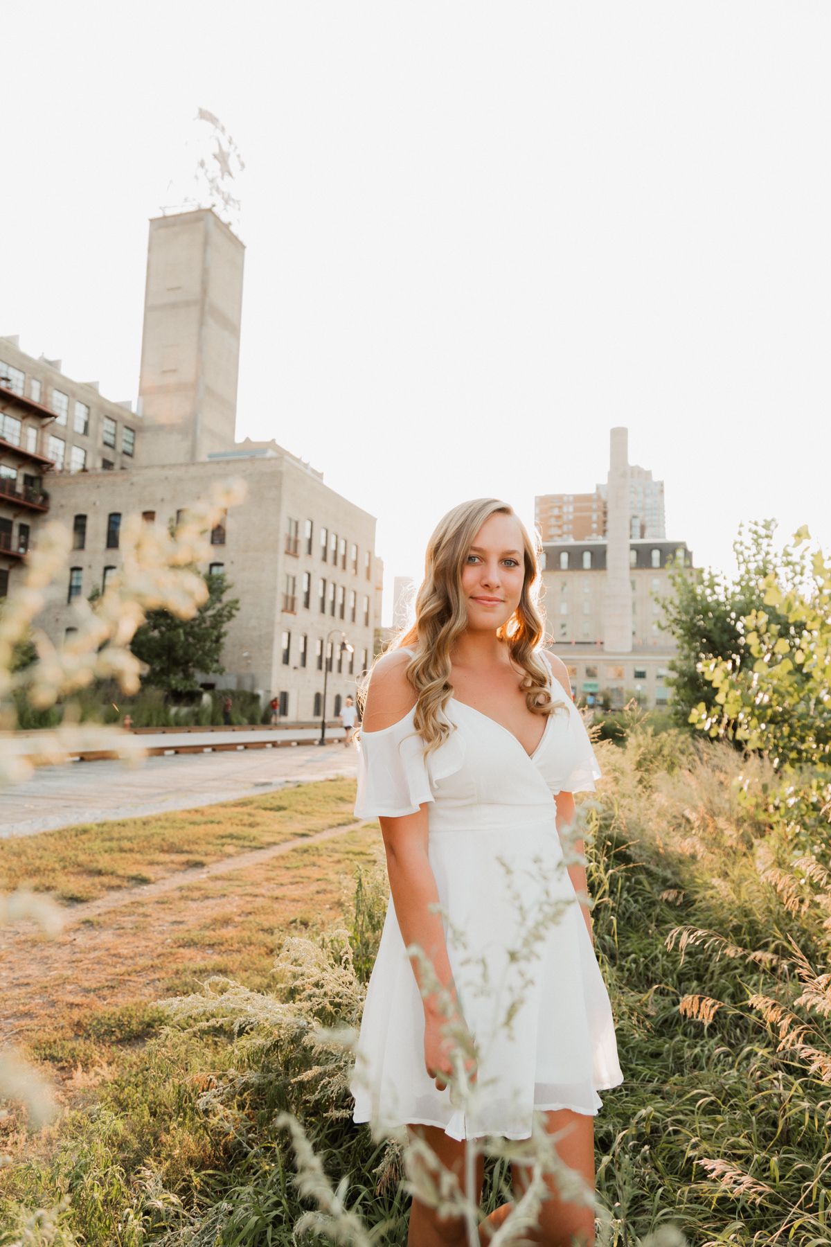 Young blonde woman in white off-shoulder dress standing in urban green space at golden hour.