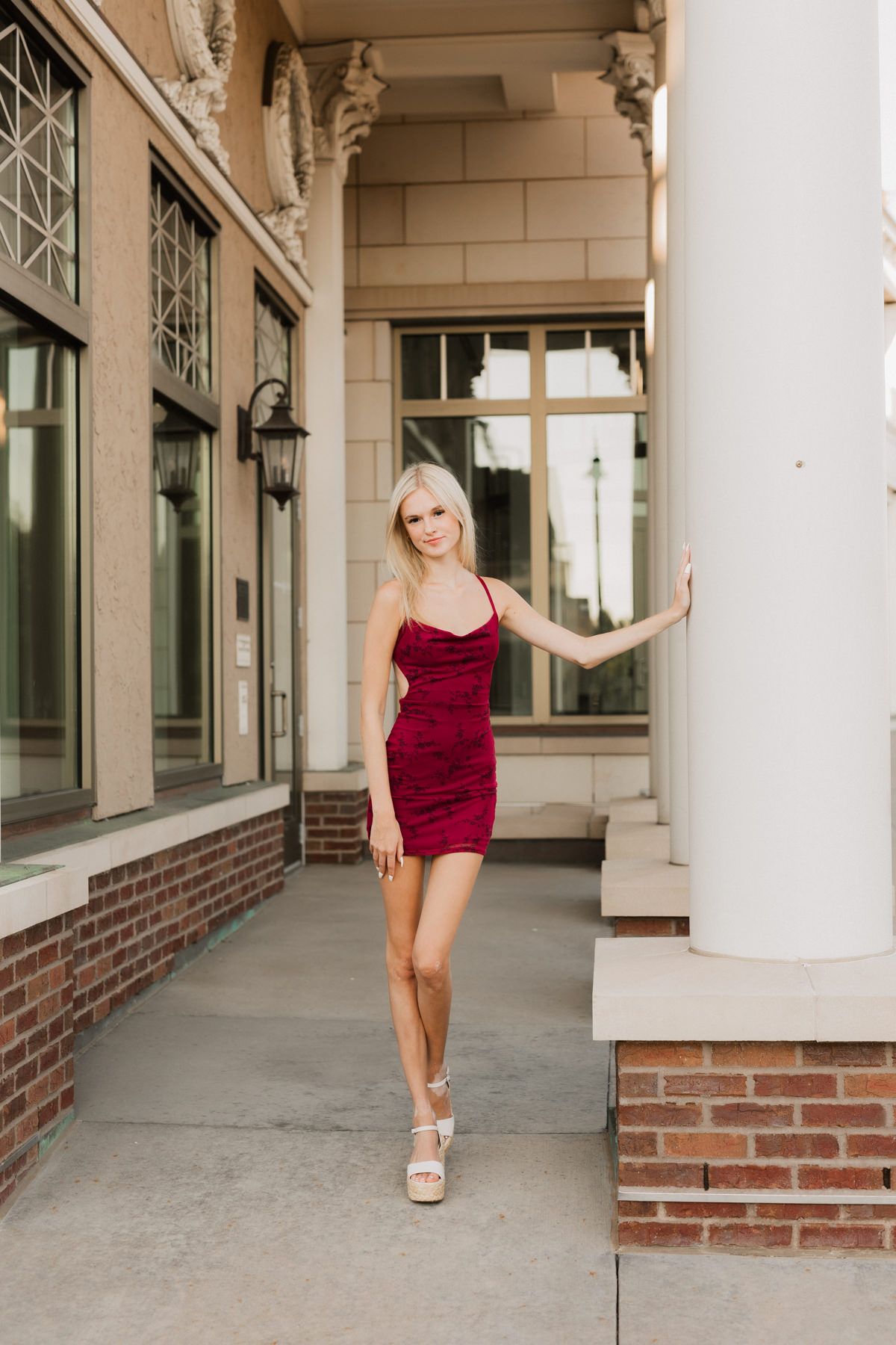 Blonde woman in red mini dress posing by white column outside historic brick building.