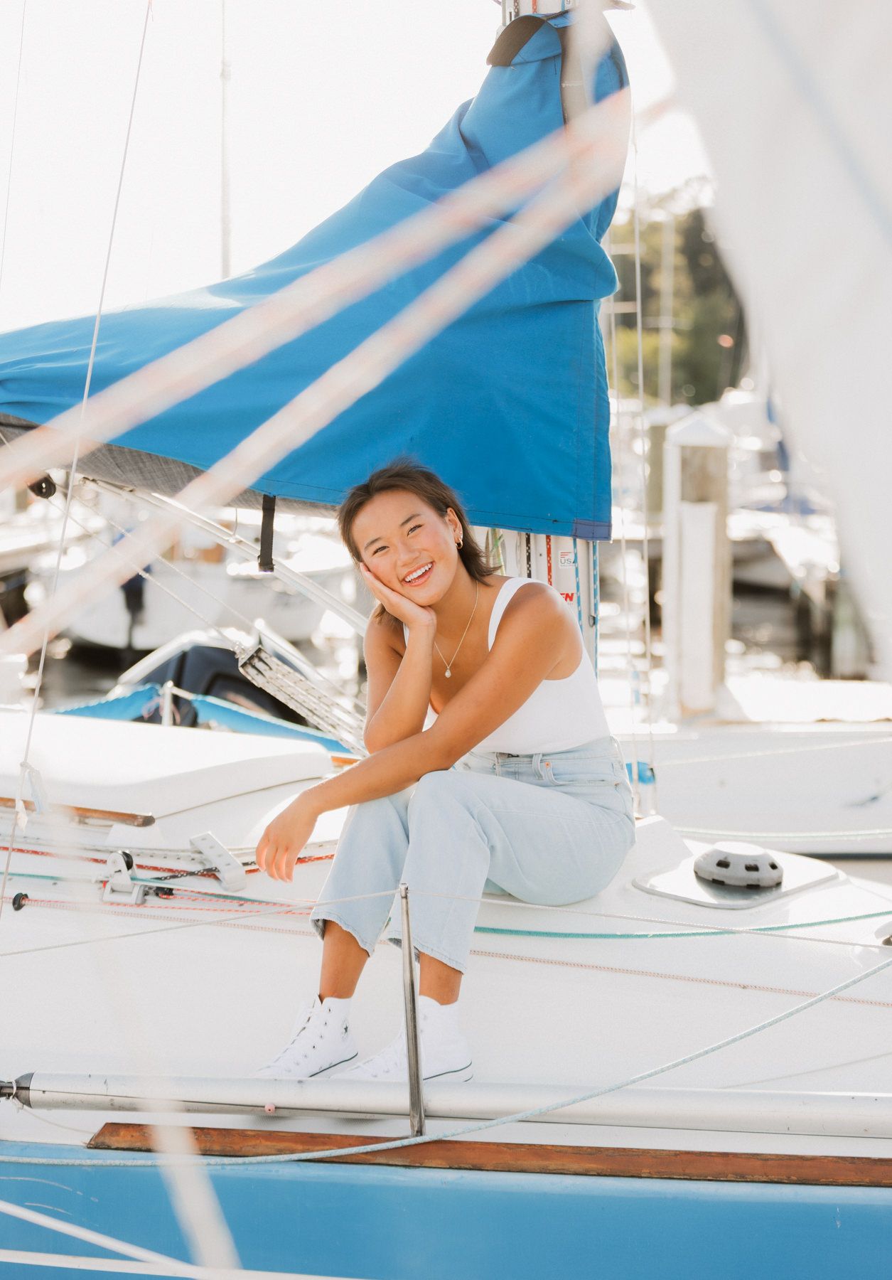 Young woman in white tank top and jeans smiling on a sailboat at a sunny marina.