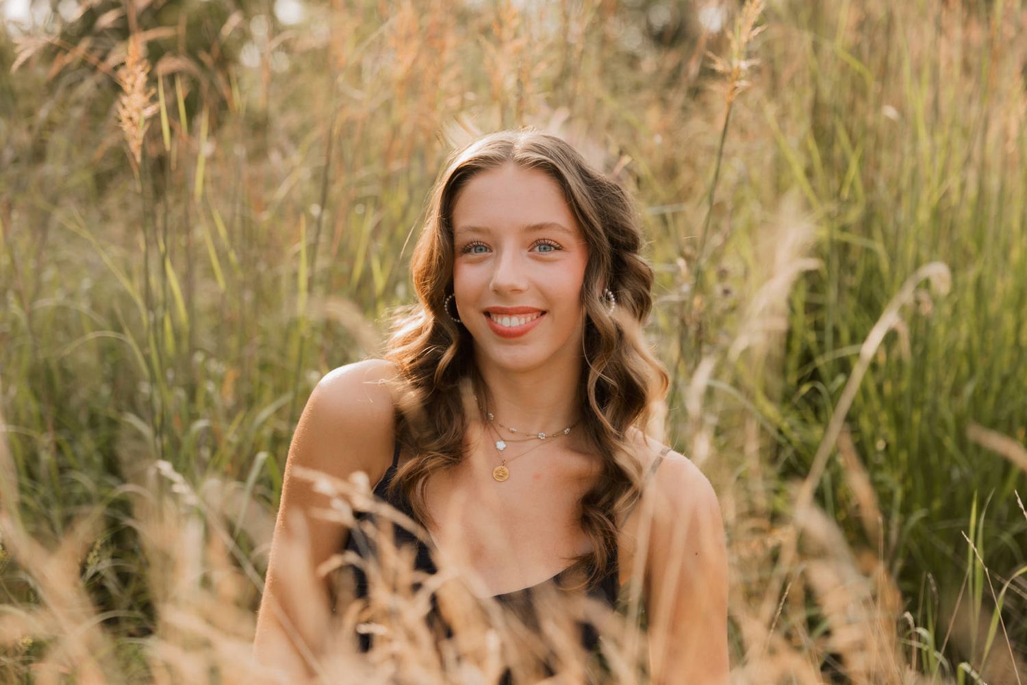 Smiling young woman with wavy hair wearing a necklace surrounded by tall golden grasses outdoors.