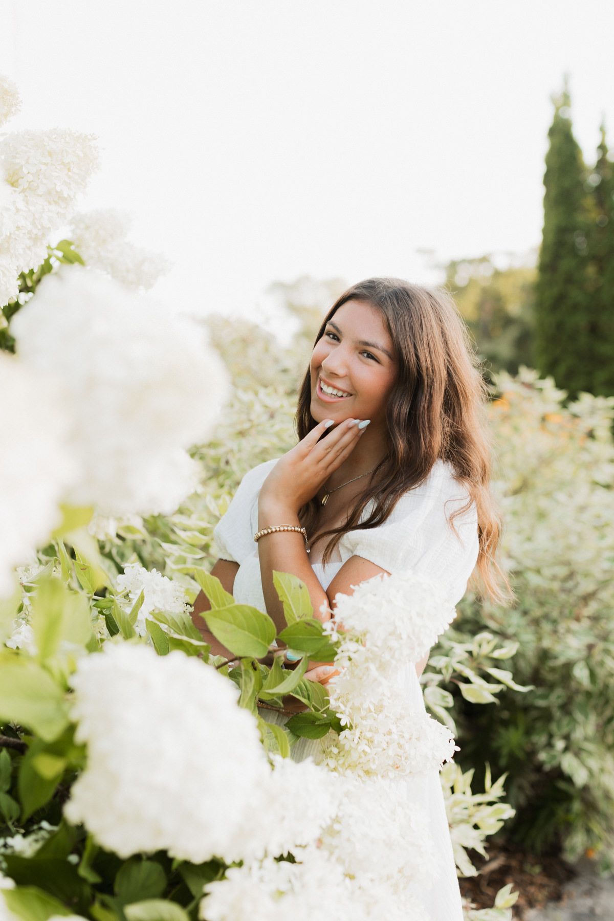 Smiling young woman in white dress surrounded by large white hydrangea blooms in garden.