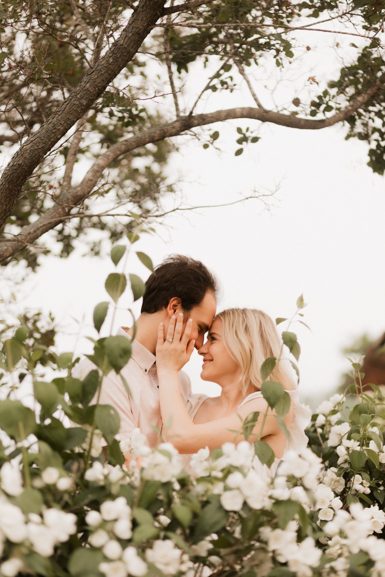 Couple sharing a romantic moment surrounded by white blooming flowers and lush green trees outdoors.
