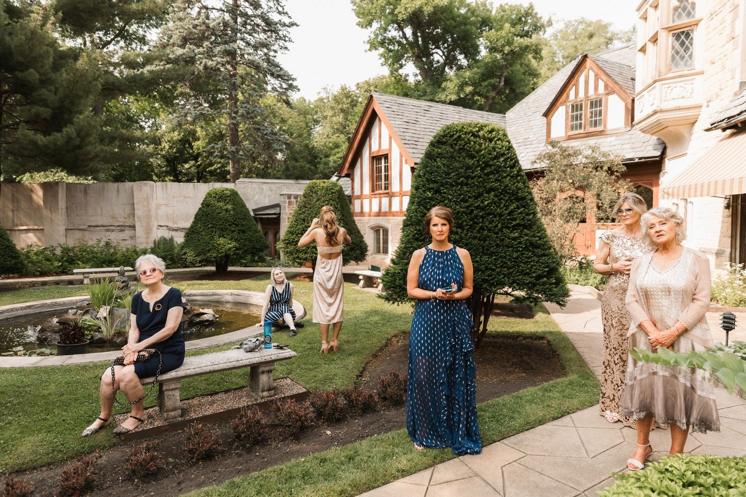 Women in elegant dresses gather in a lush garden beside a Tudor-style estate during an outdoor wedding celebration.