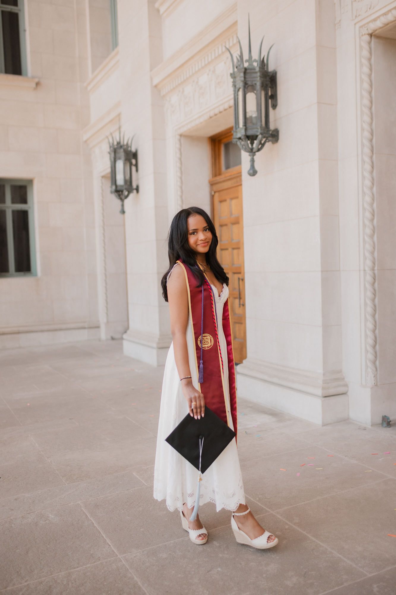 Young woman in white dress with red graduation stole holds cap, posing outdoors at university building.