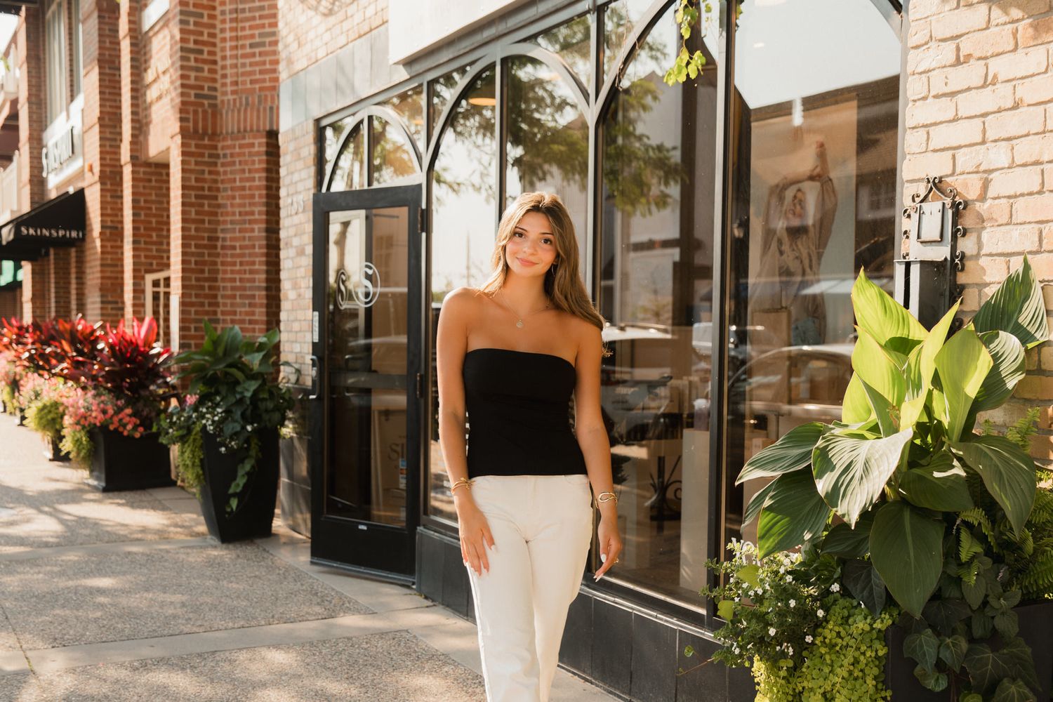 Woman in black strapless top and white pants posing outside a boutique shop on a sunny day.