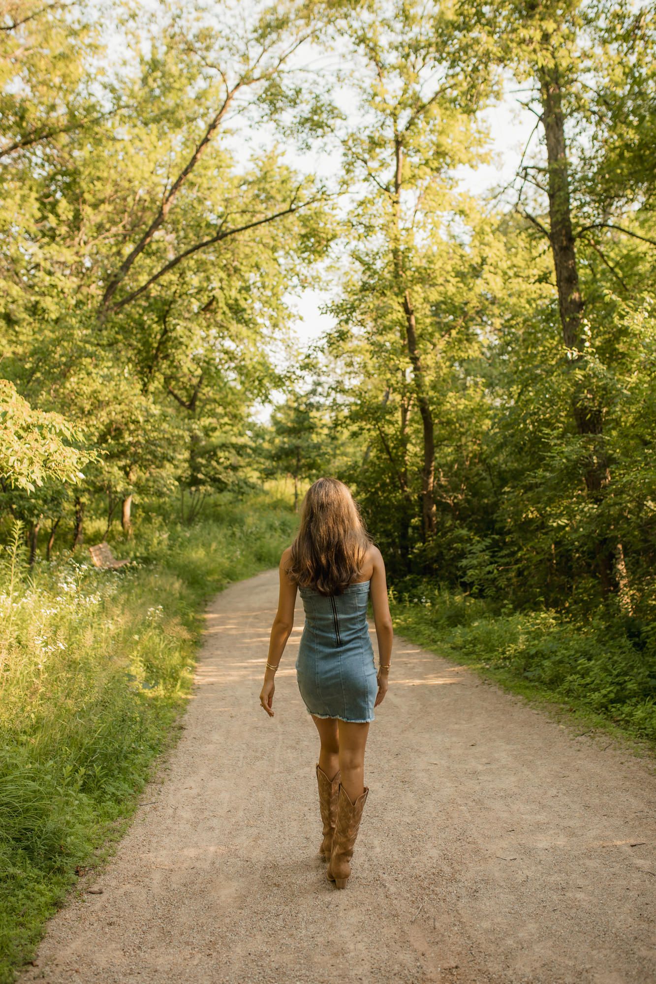 Woman in denim dress and cowboy boots walking down a scenic dirt path surrounded by lush green trees in golden sunlight.