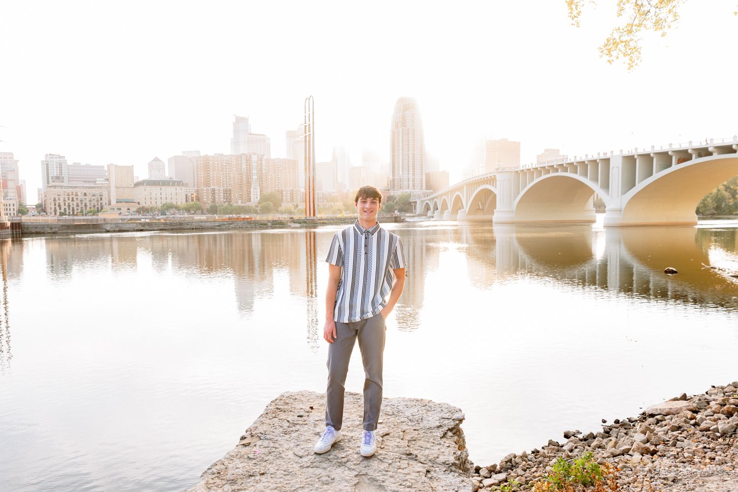 Young couple walking along riverbank at sunset with Minneapolis skyline and bridge visible in background.