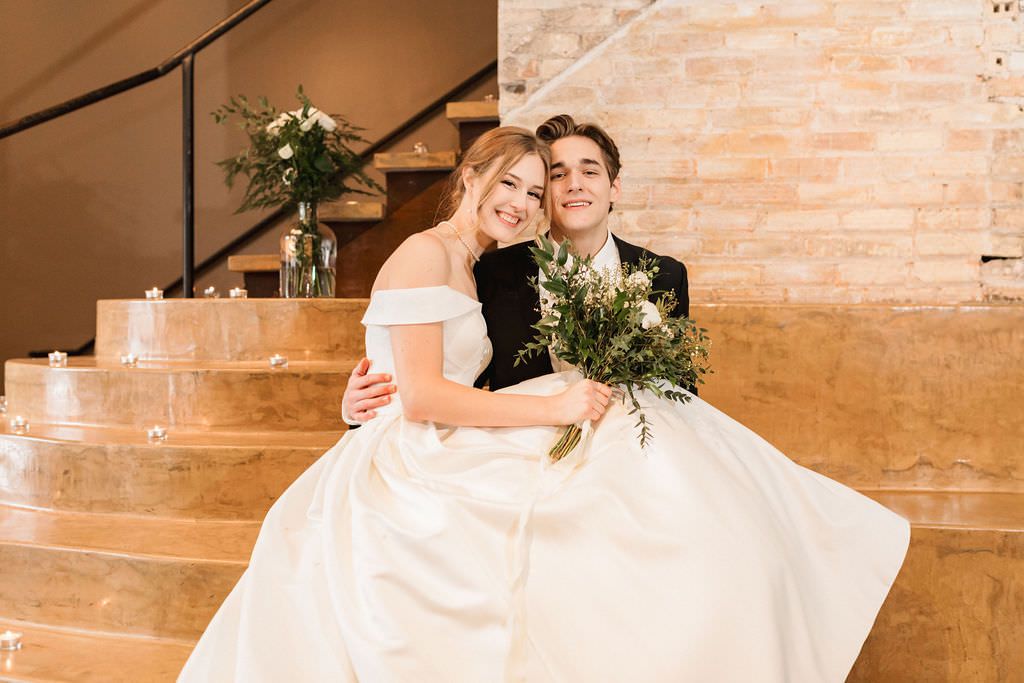 A wedding couple embraces romantically against exposed brick walls with glowing candles on the floor.