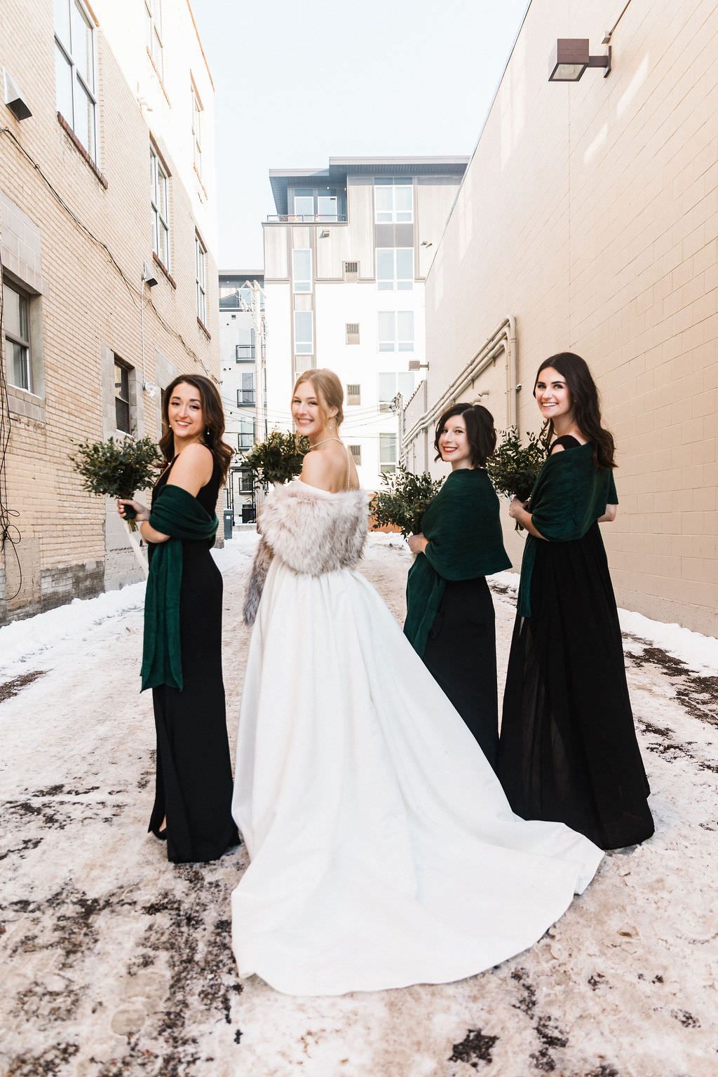 Bride in white gown poses with bridesmaids in dark green dresses against urban winter backdrop.