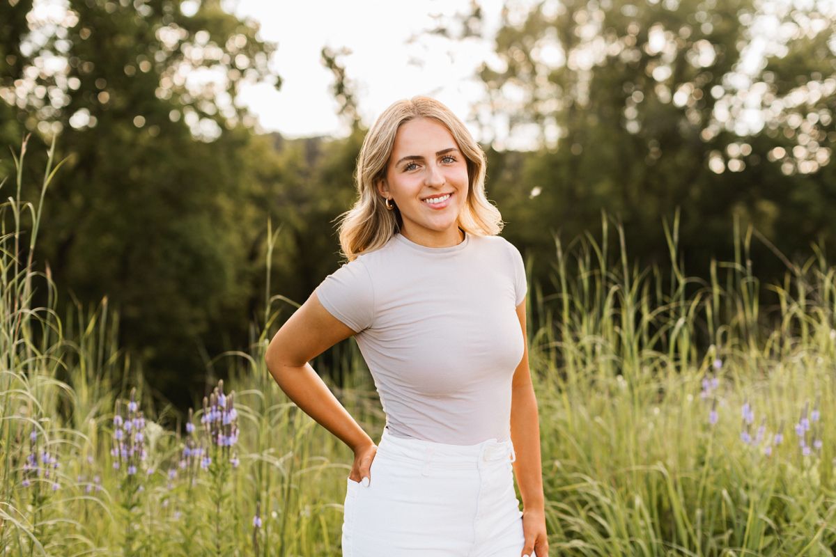 Someone poses in a casual white outfit amid tall grass and wildflowers during golden hour.