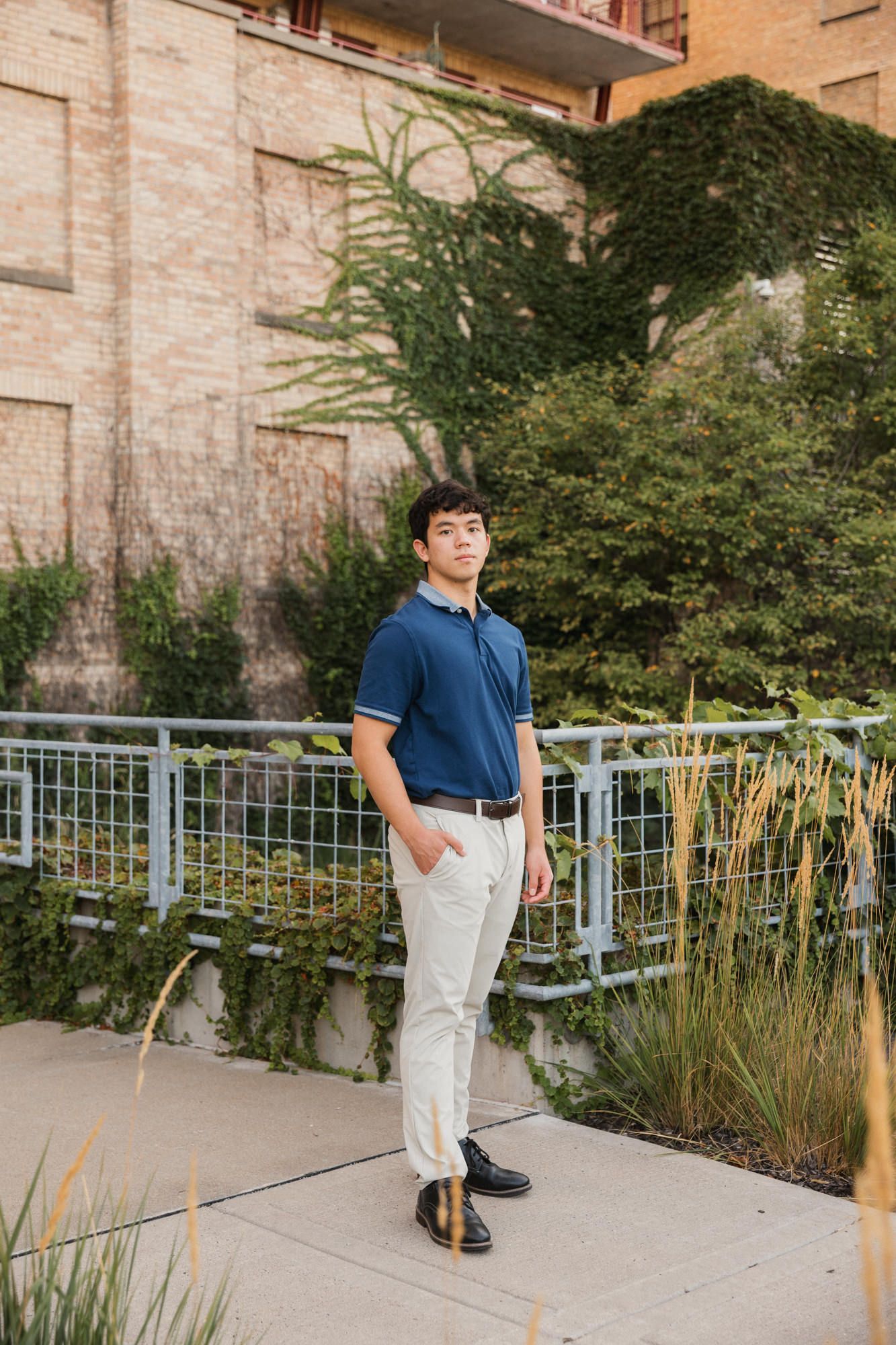Young man in blue polo shirt and cream pants standing on urban walkway with brick building and greenery.