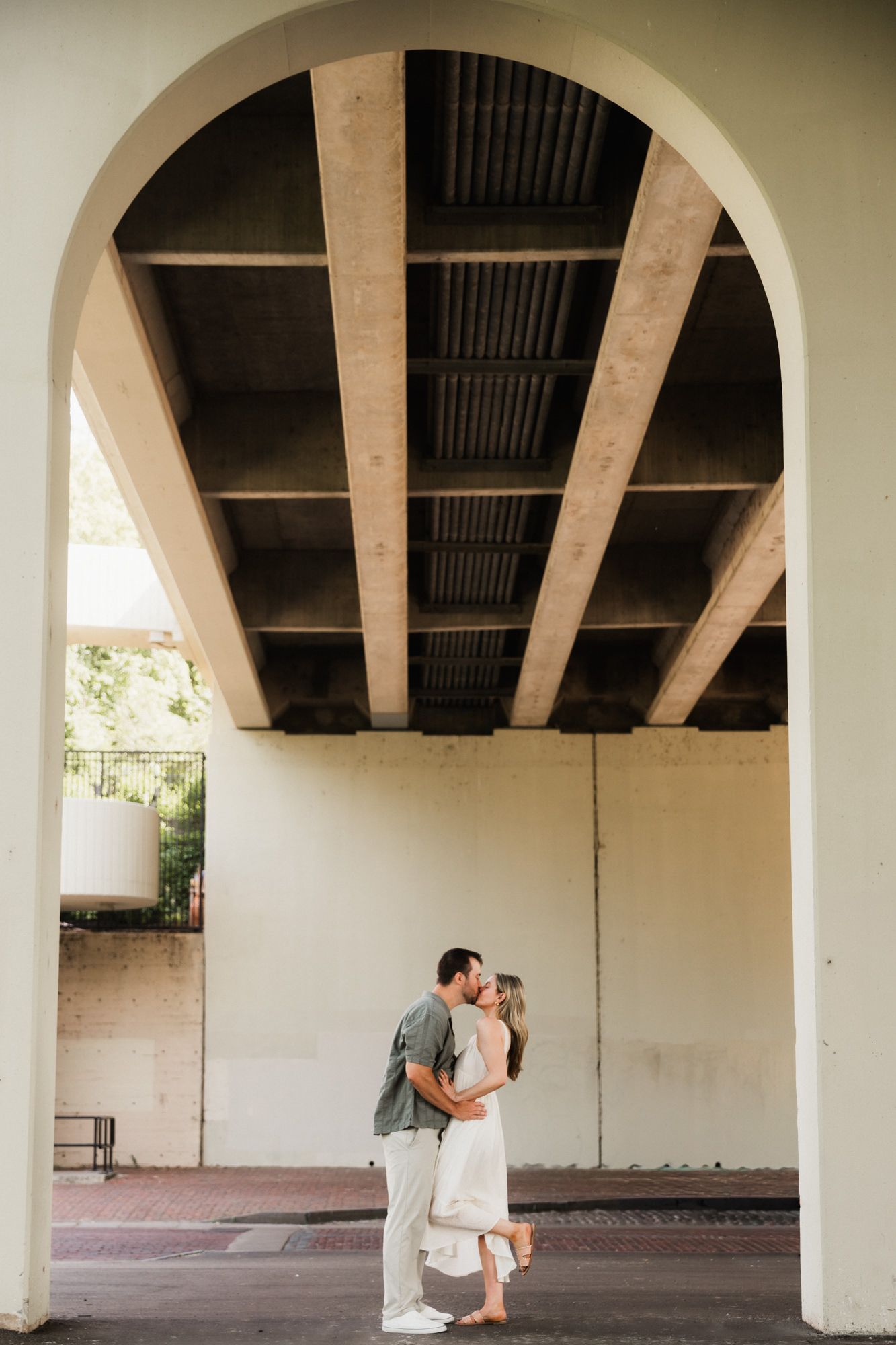 Couple sharing a romantic kiss under a concrete arch bridge, man in green shirt and woman in white dress.