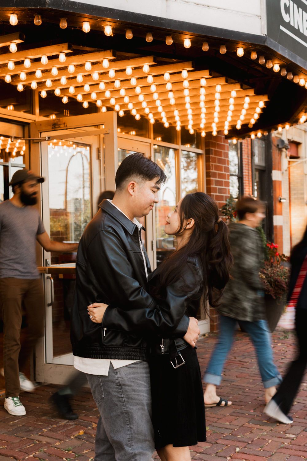 A couple embraces outside a theater entrance with glowing marquee lights and brick walls in an evening setting.