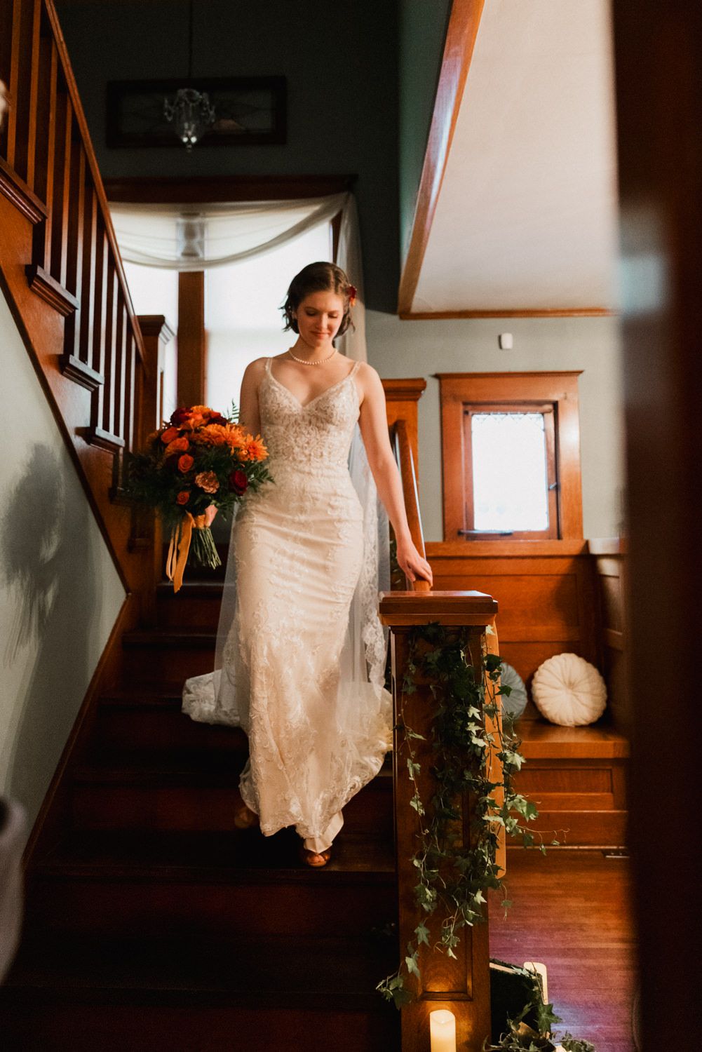 A bride in a lace wedding gown descends wooden stairs while holding a vibrant orange bouquet.