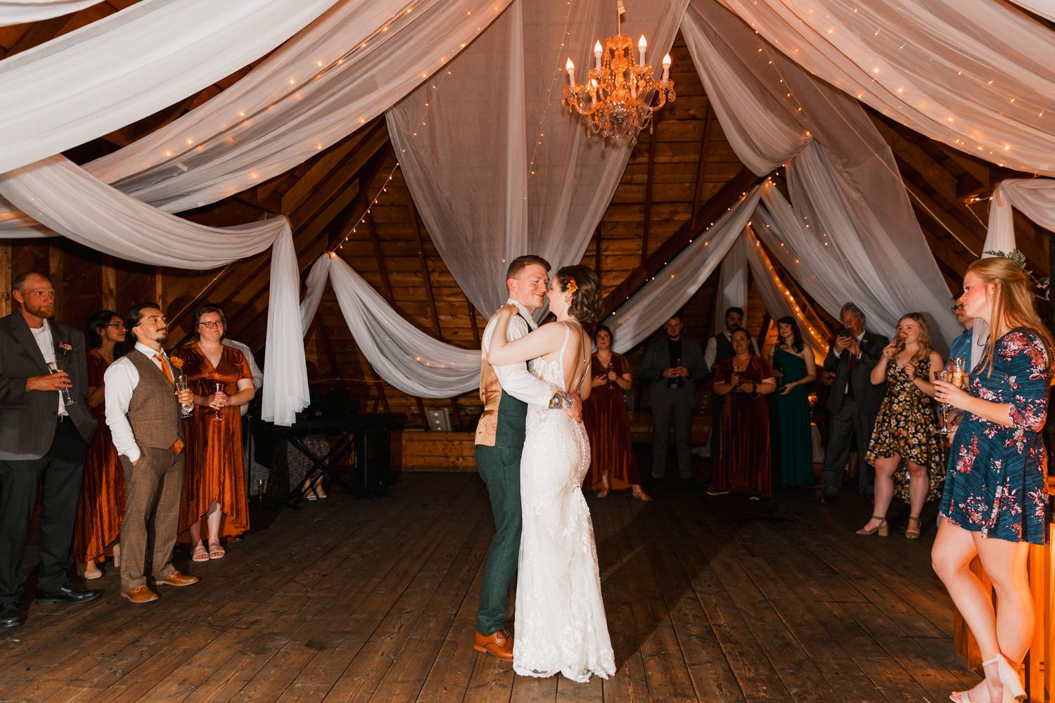 A couple shares their first dance at a rustic barn wedding reception under draped fabric and warm lighting.