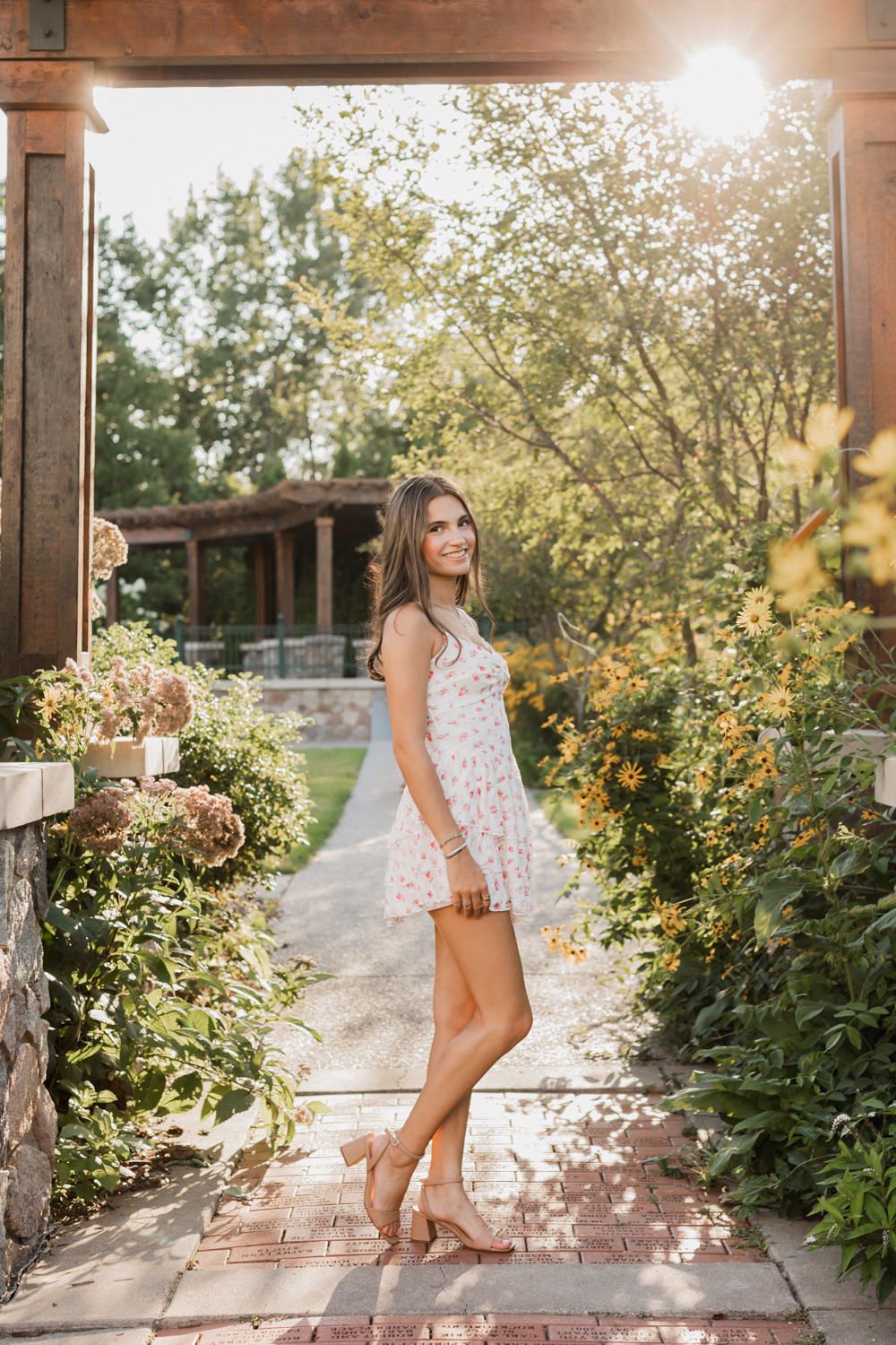 A person in a white floral dress poses on a rustic porch surrounded by blooming flowers and sunlit greenery.