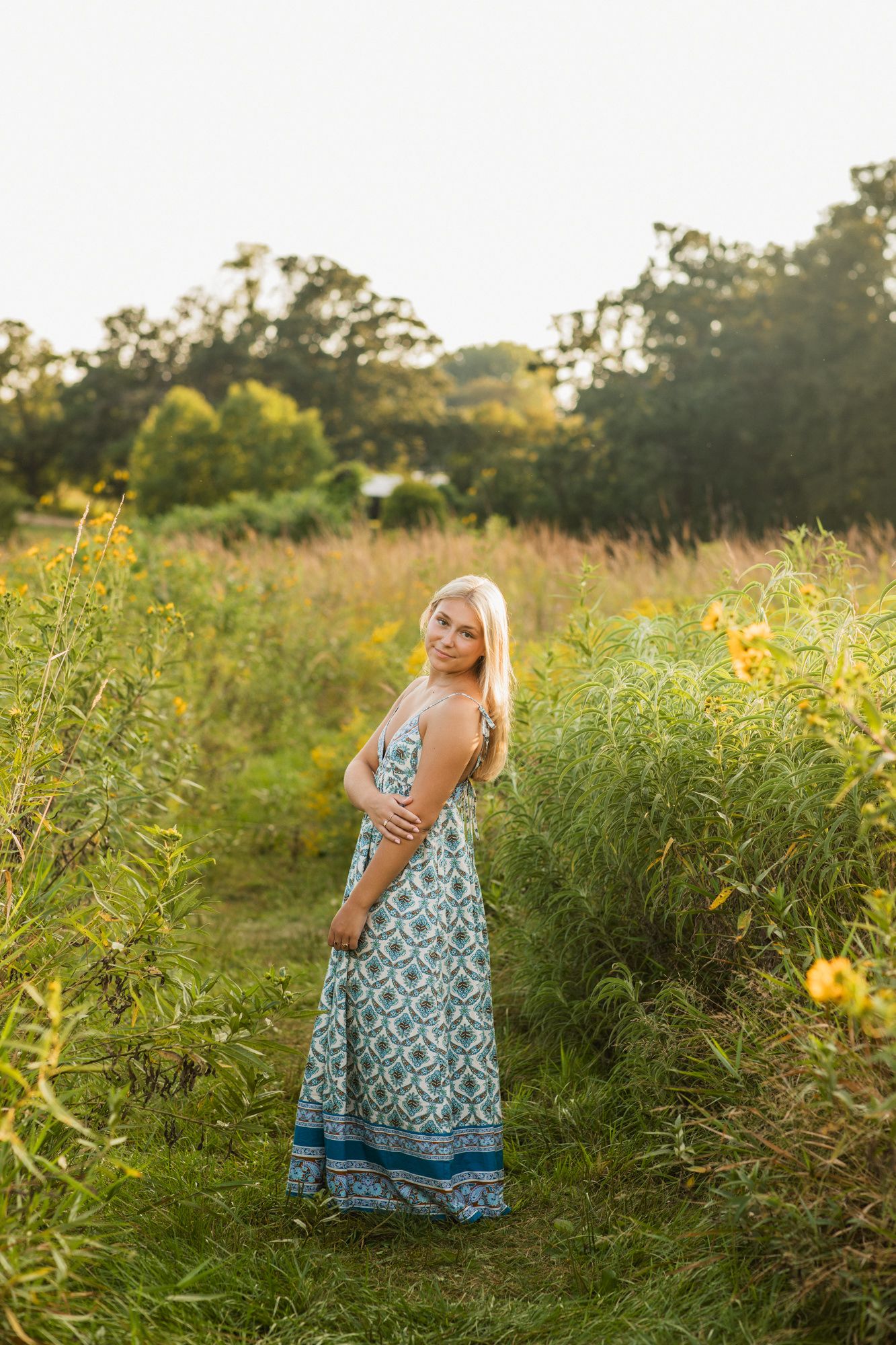 Blonde woman in a blue patterned maxi dress standing in a lush green field with yellow wildflowers at golden hour.