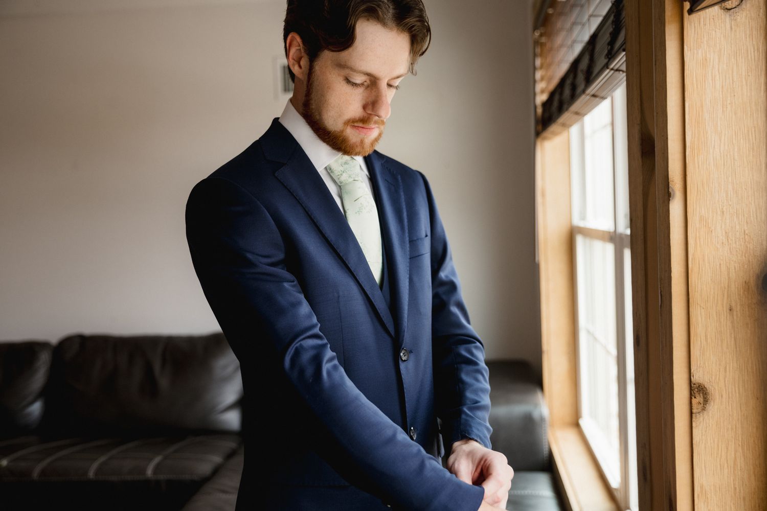 A groom in a navy blue suit adjusts his cufflinks while standing near a window in soft natural light.