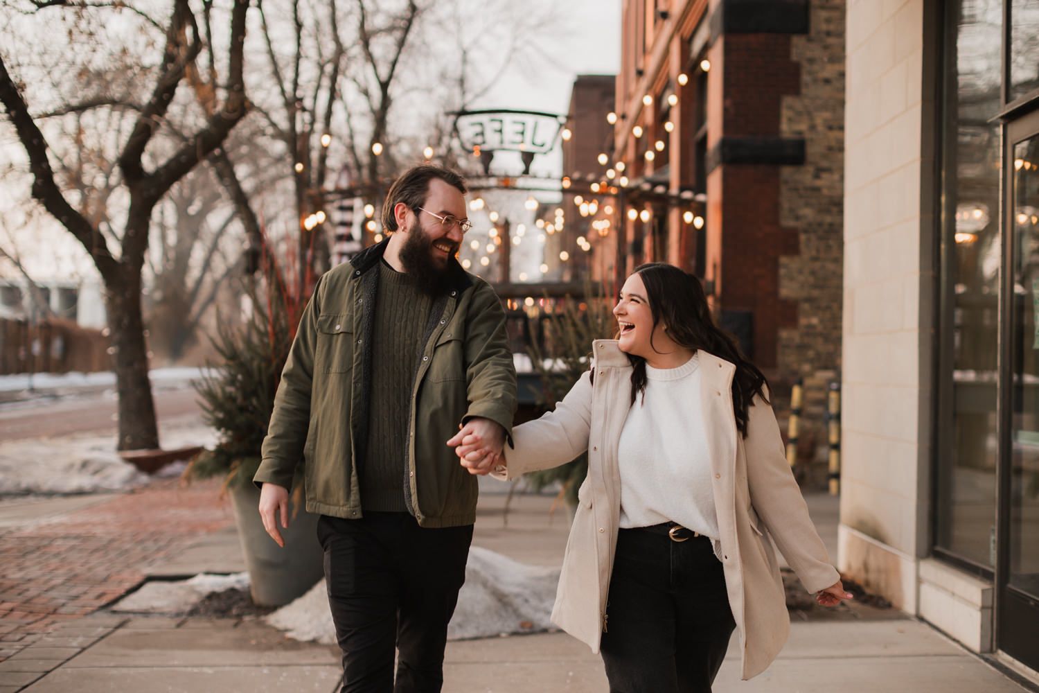 Couple walking together along a brick sidewalk with storefronts and string lights visible.