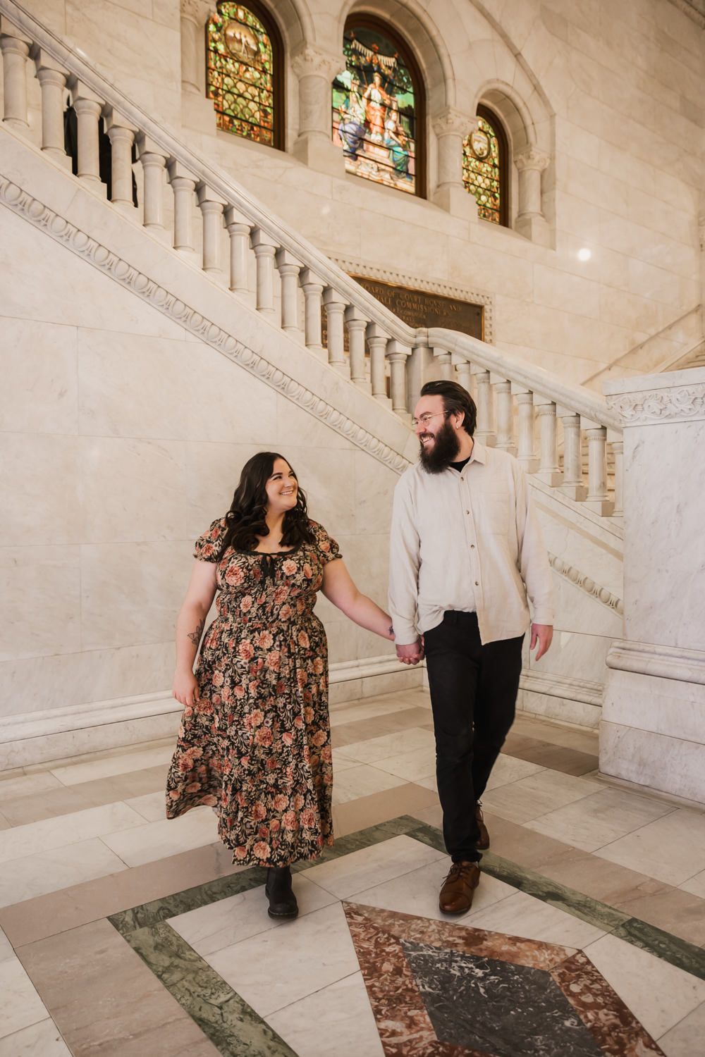 A couple walks hand in hand on marble stairs with stained glass windows in an elegant historic building interior.