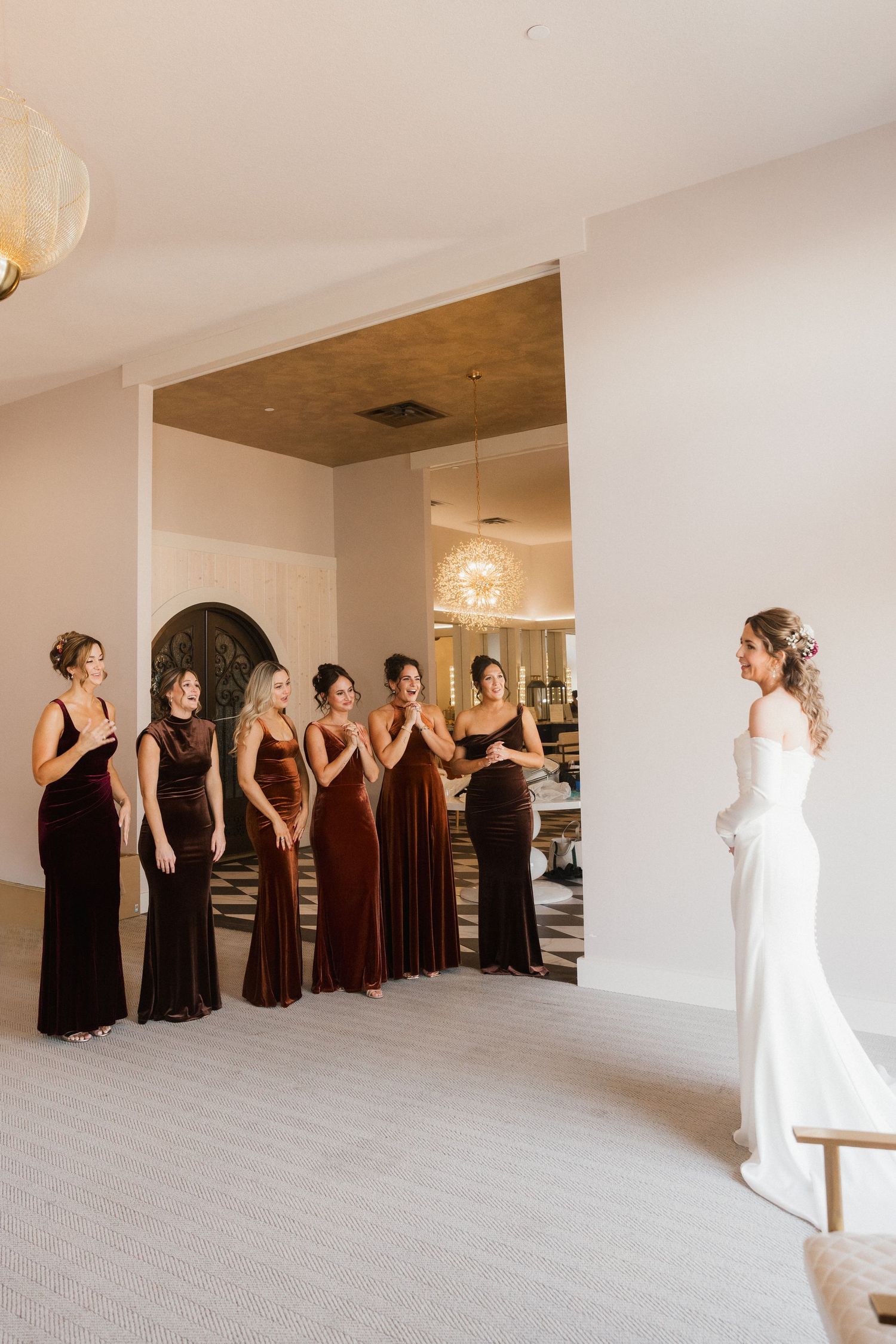Bridesmaids in rich velvet dresses clasp their hands in joy during an emotional first look with the bride in an elegant white gown.