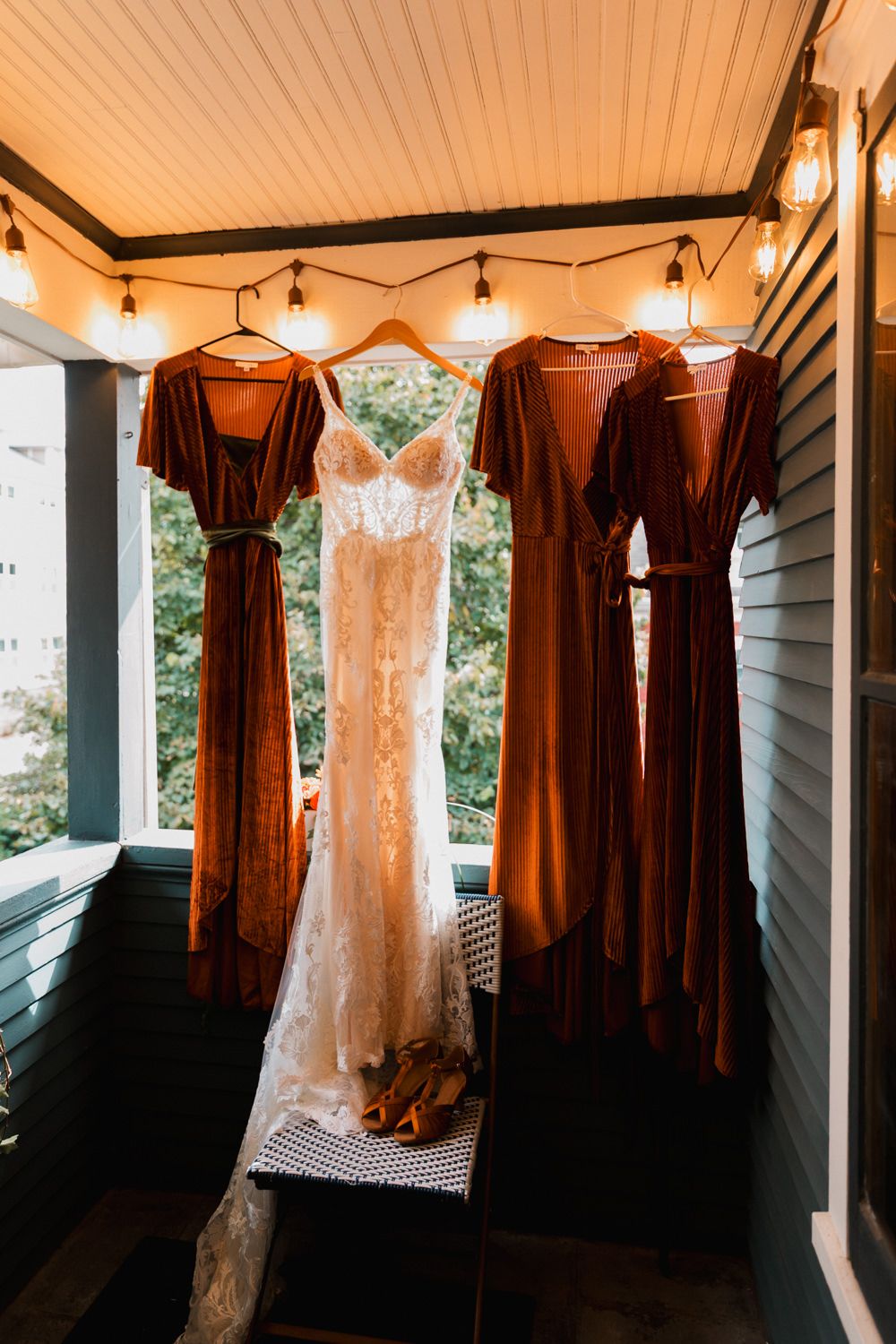 Wedding dresses and burgundy bridesmaid gowns hanging on display against a white wall with string lights.