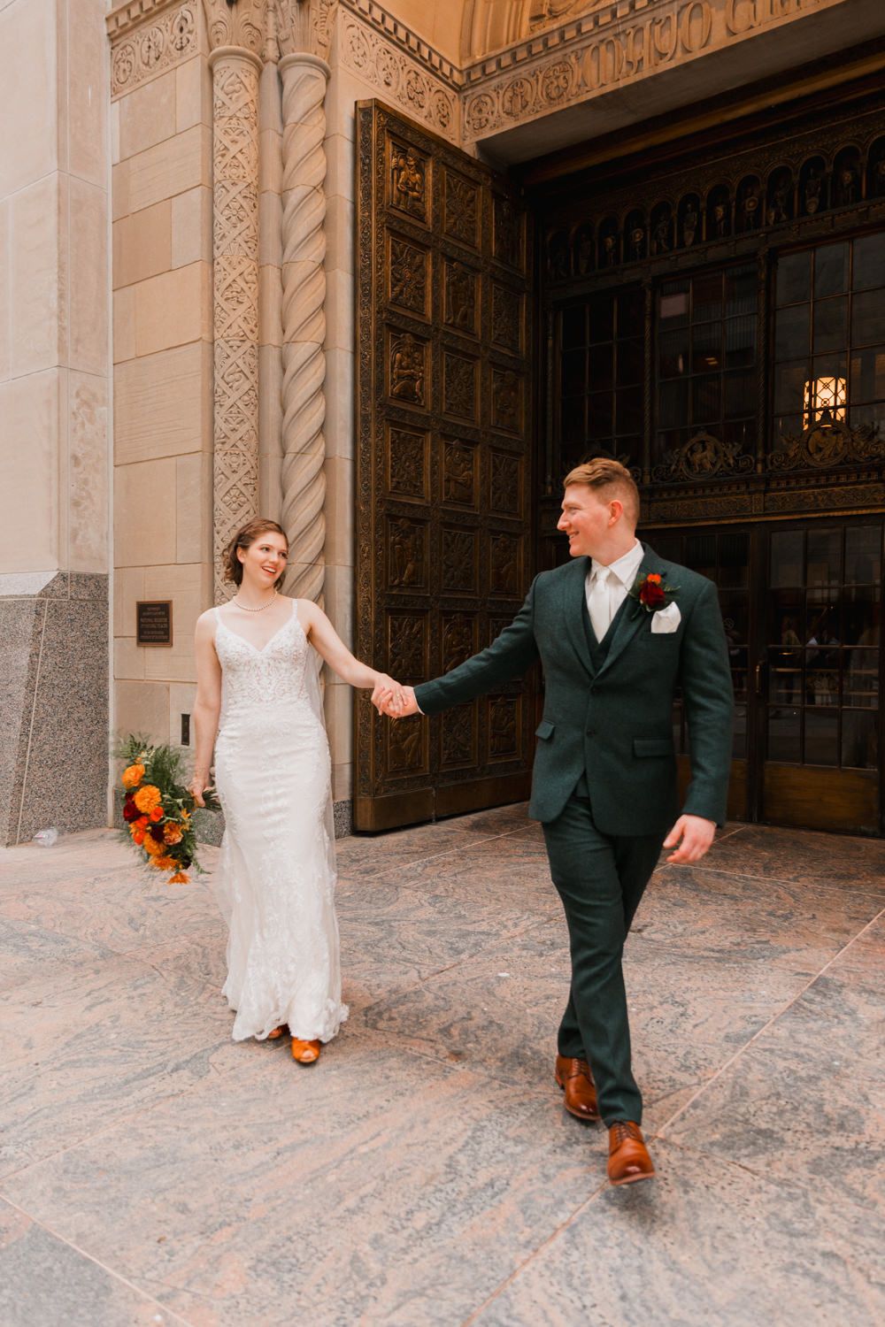 Couple in wedding attire playfully dance and twirl in an ornate marble hallway with decorative stone walls.