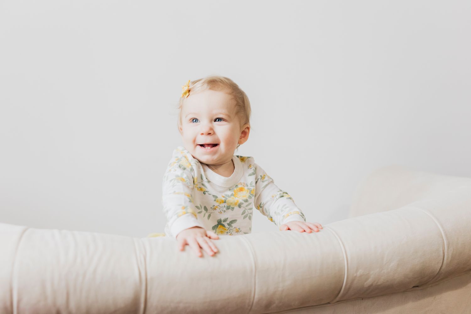 A baby in floral clothing smiles while sitting on a white couch.