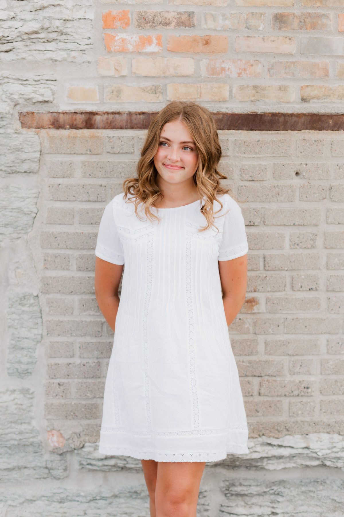 Young woman in white short-sleeve dress posing confidently against a rustic brick wall.