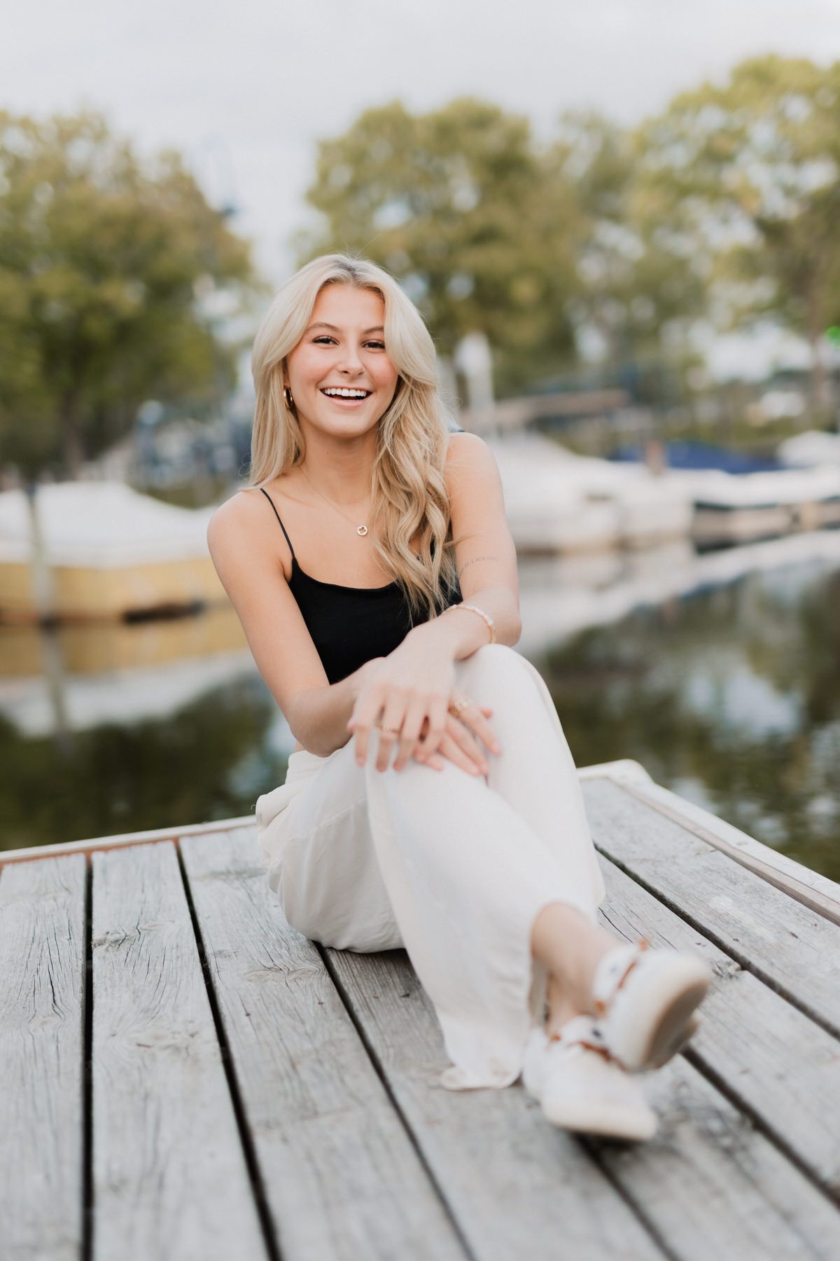 Laughing blonde woman sitting on a wooden dock near boats, wearing a black top and white pants.