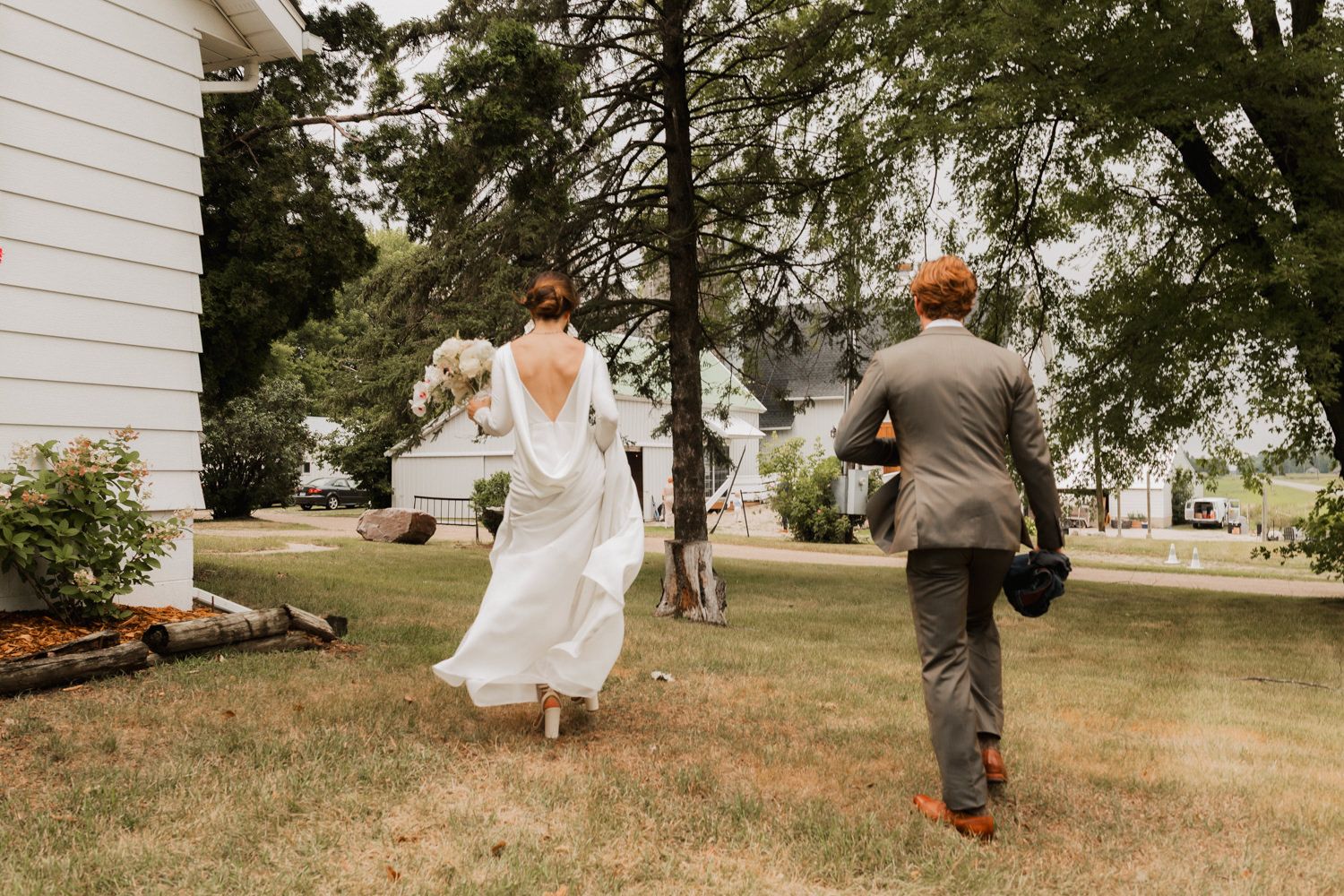 A couple in wedding attire runs playfully across a lawn near a white house surrounded by trees.