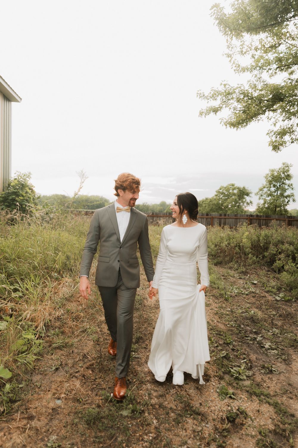 A couple in wedding attire walks together on a rustic hillside path during an outdoor photo session.