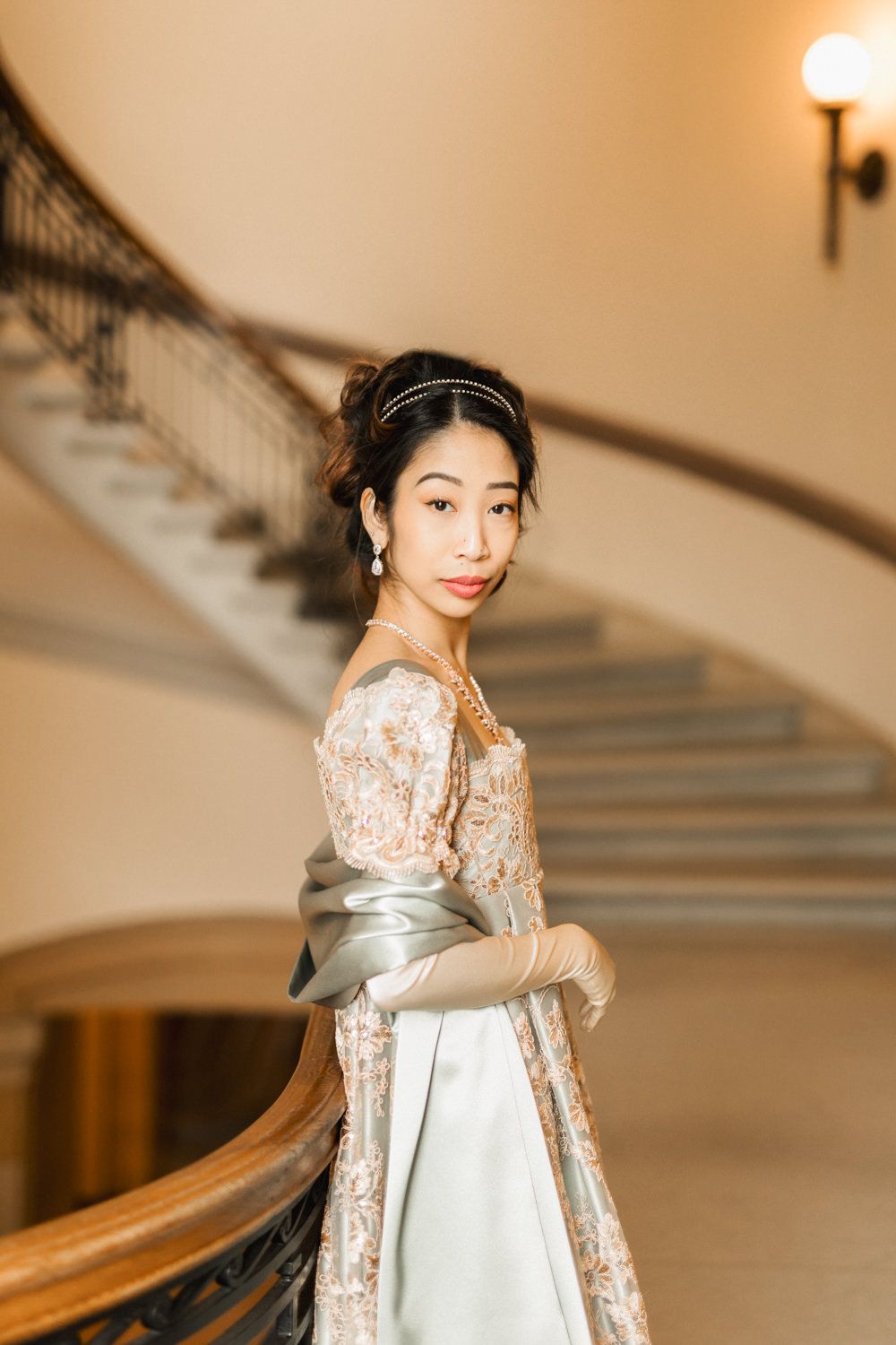 Young Asian woman in elegant floral gown with satin gloves poses gracefully on ornate curved staircase in grand interior.