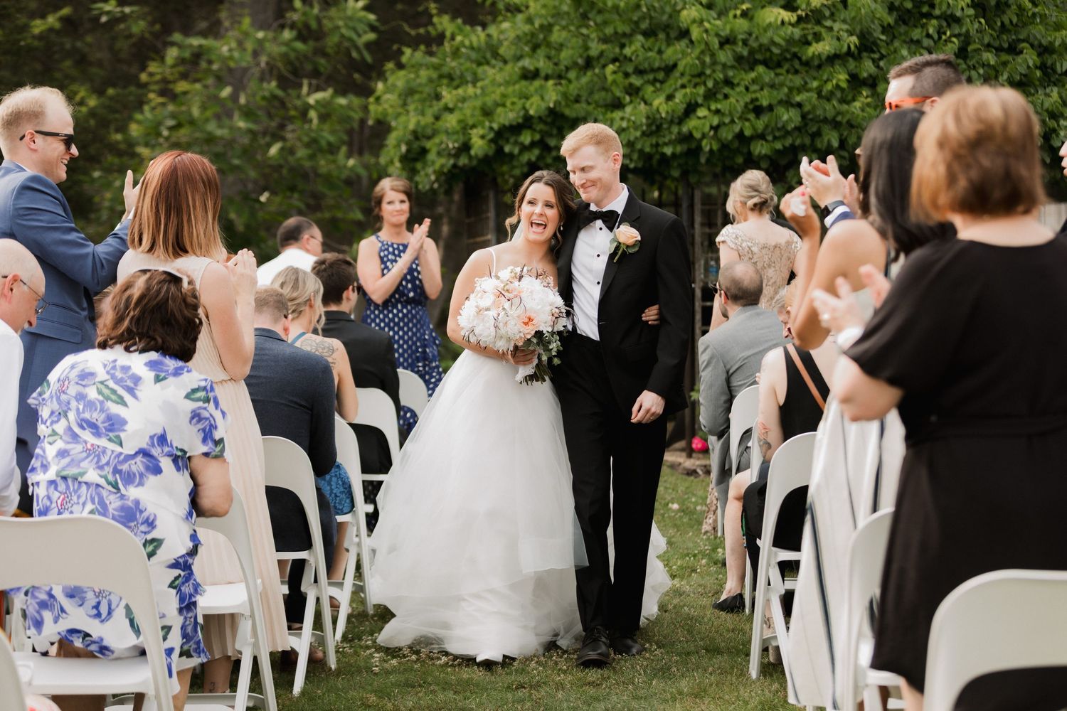 Bride and groom walk down the aisle at an outdoor wedding ceremony, guests applauding in celebration.