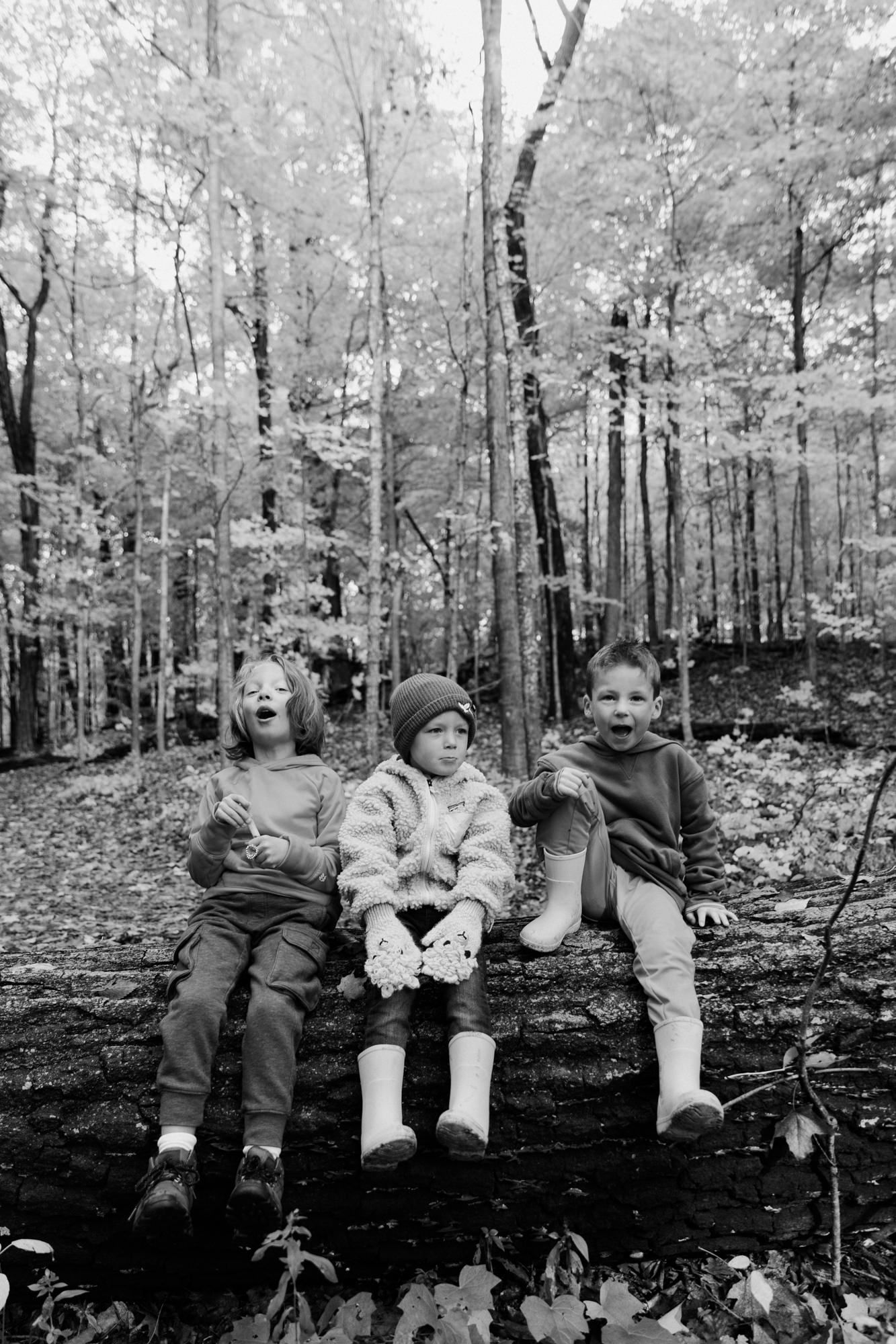 Three children sit on a log in an autumn forest, smiling and posing for a black and white photo.