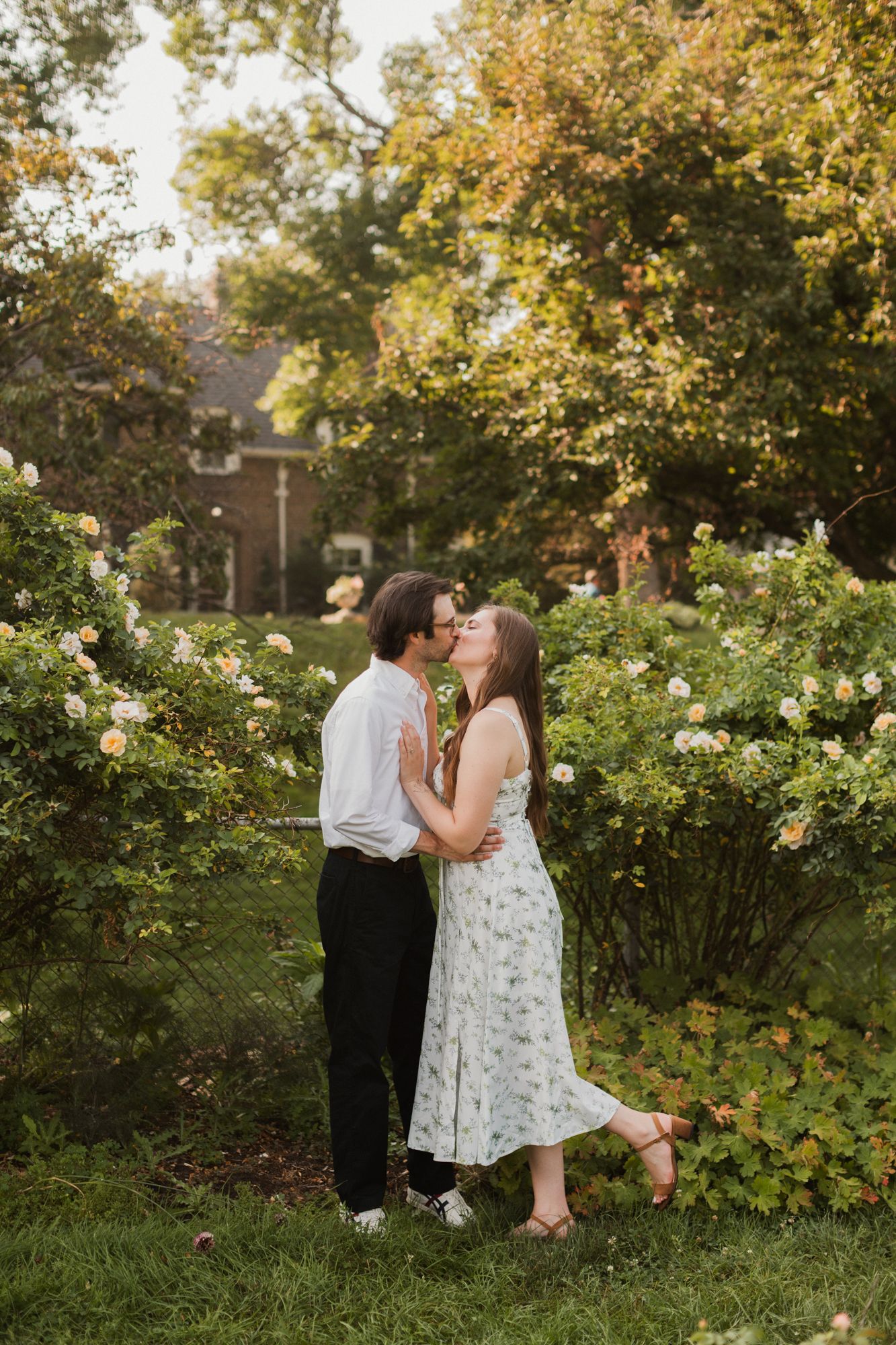 A couple shares a romantic kiss surrounded by blooming rose bushes in a lush garden setting.