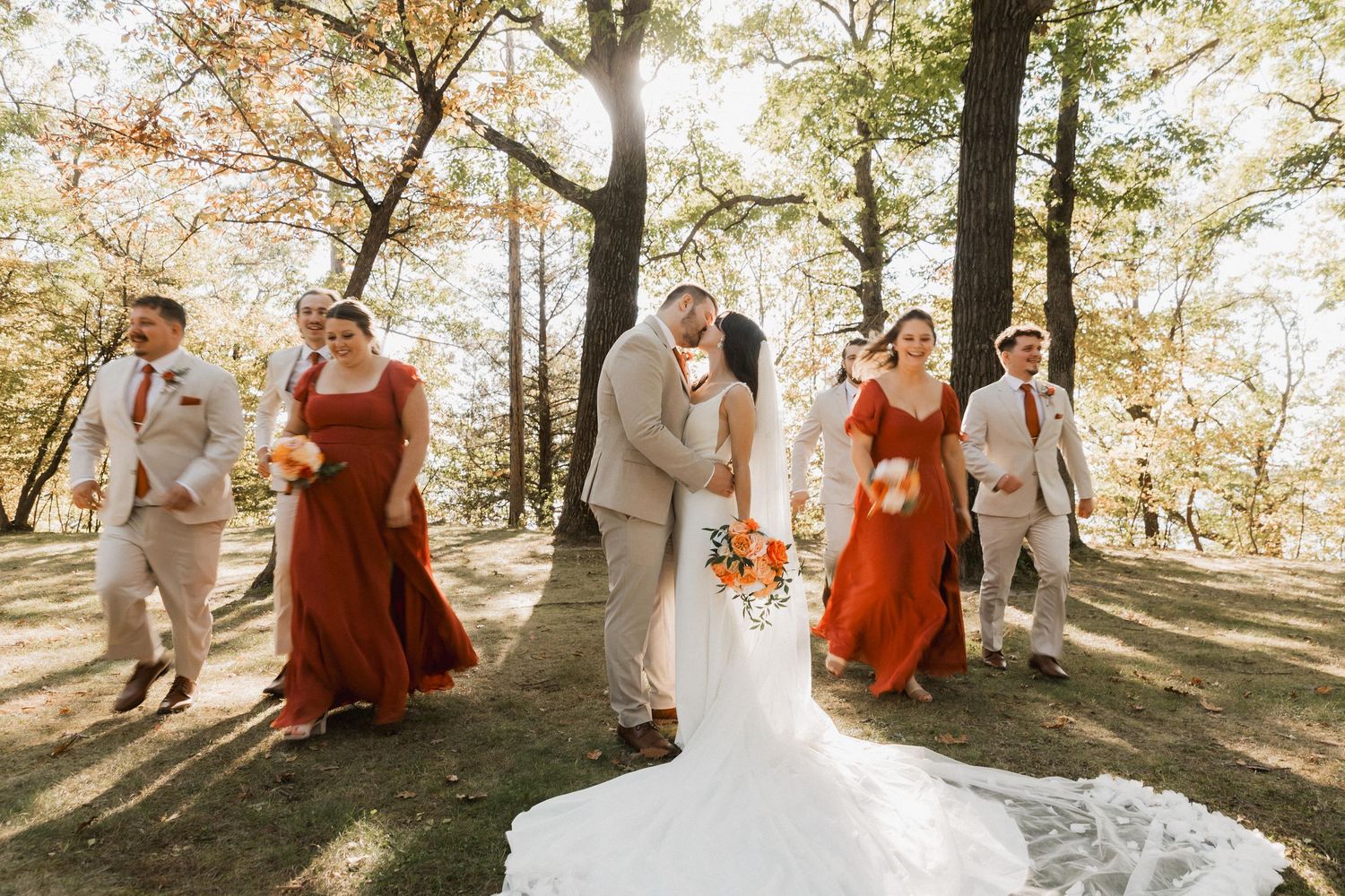 Wedding party in autumn forest wearing white and rust-colored dresses dance and celebrate in golden sunlight.