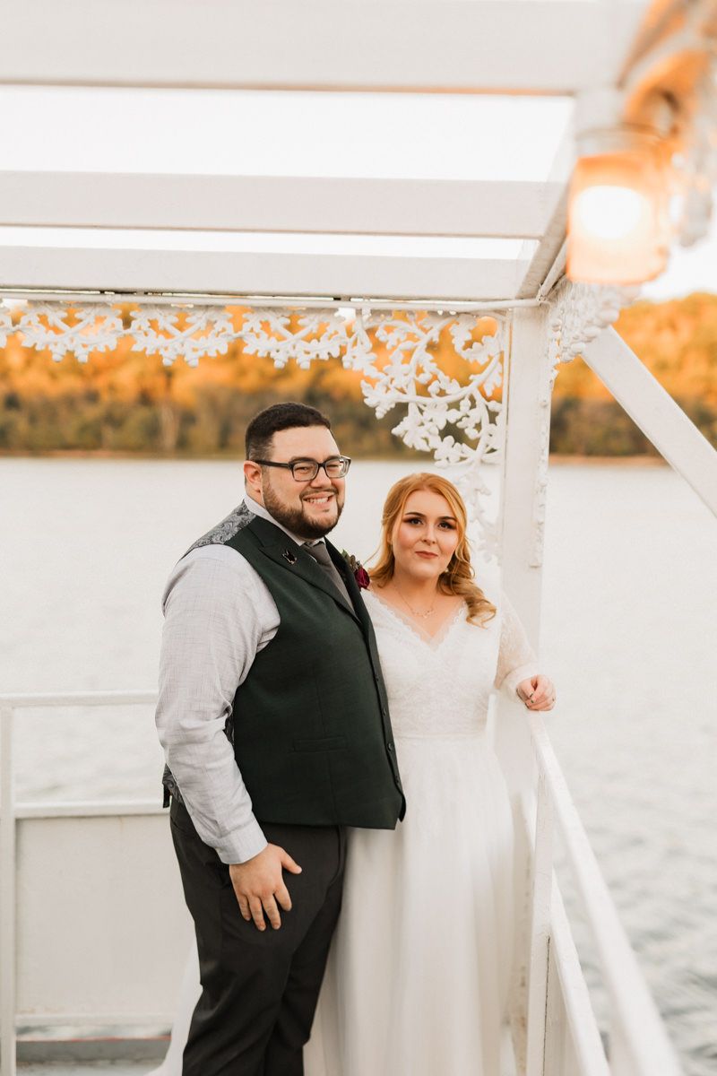 A joyful wedding couple shares a candid moment on a boat deck decorated with twinkle lights at sunset.