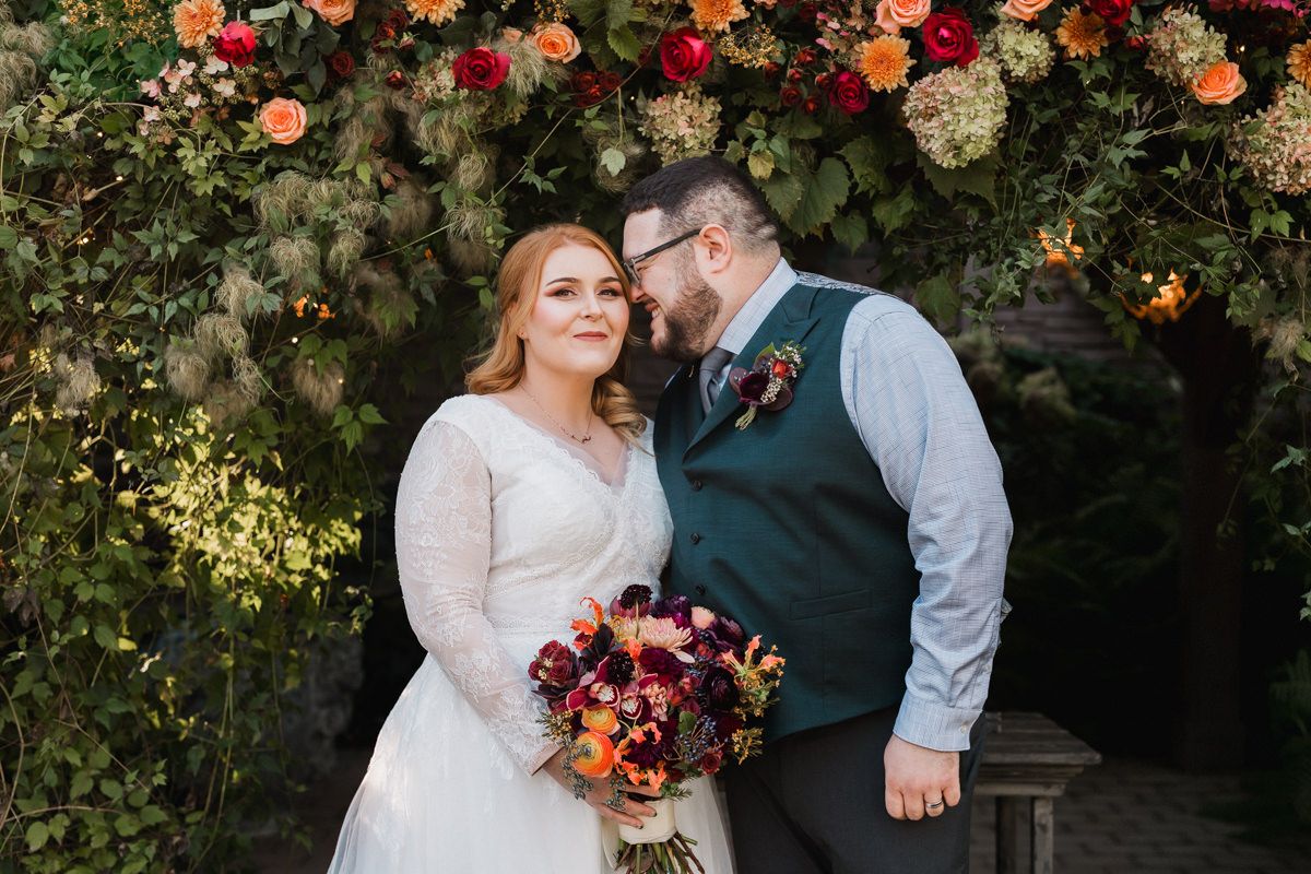 Wedding couple embracing in garden setting with fall-colored floral arrangements.